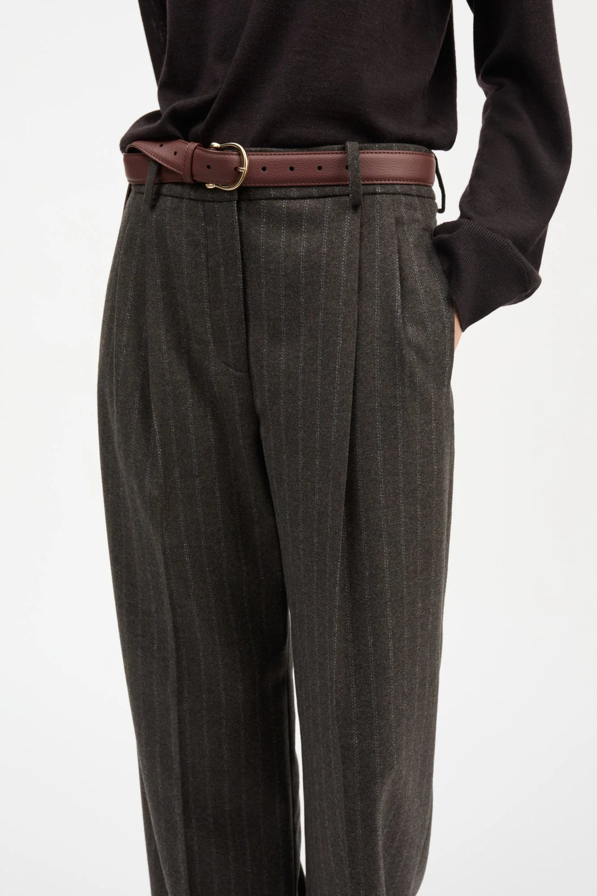 A close-up of a person wearing dark charcoal gray pinstriped trousers, a brown leather belt with a gold buckle, and a black long-sleeve top against a plain white background.