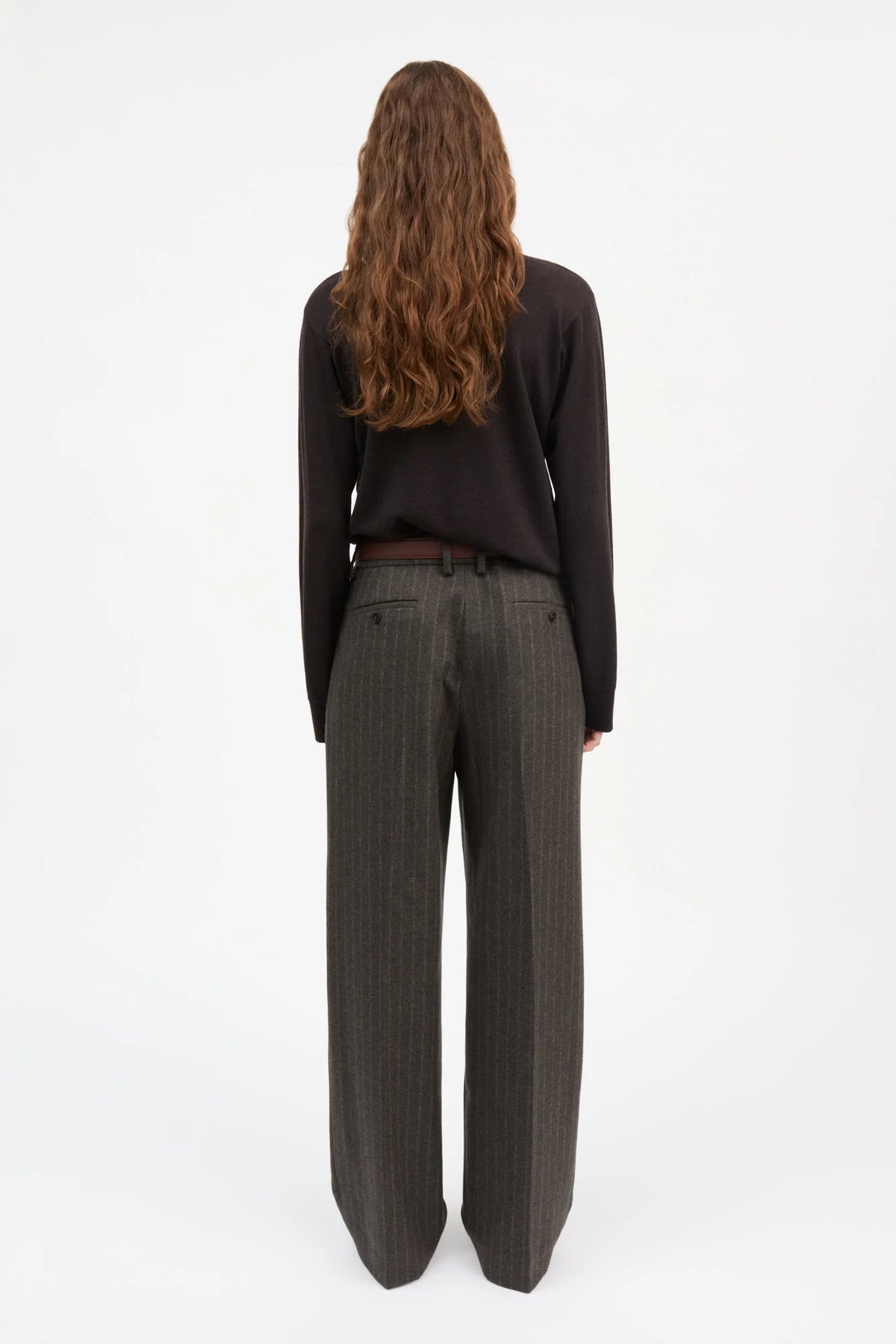 A woman viewed from behind wearing a black long-sleeve top and dark gray pinstripe wide-leg trousers, standing against a plain white studio background.