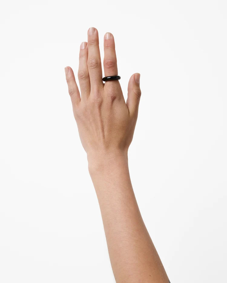A minimalist black ring worn on the ring finger of a hand, shown against a plain white background.