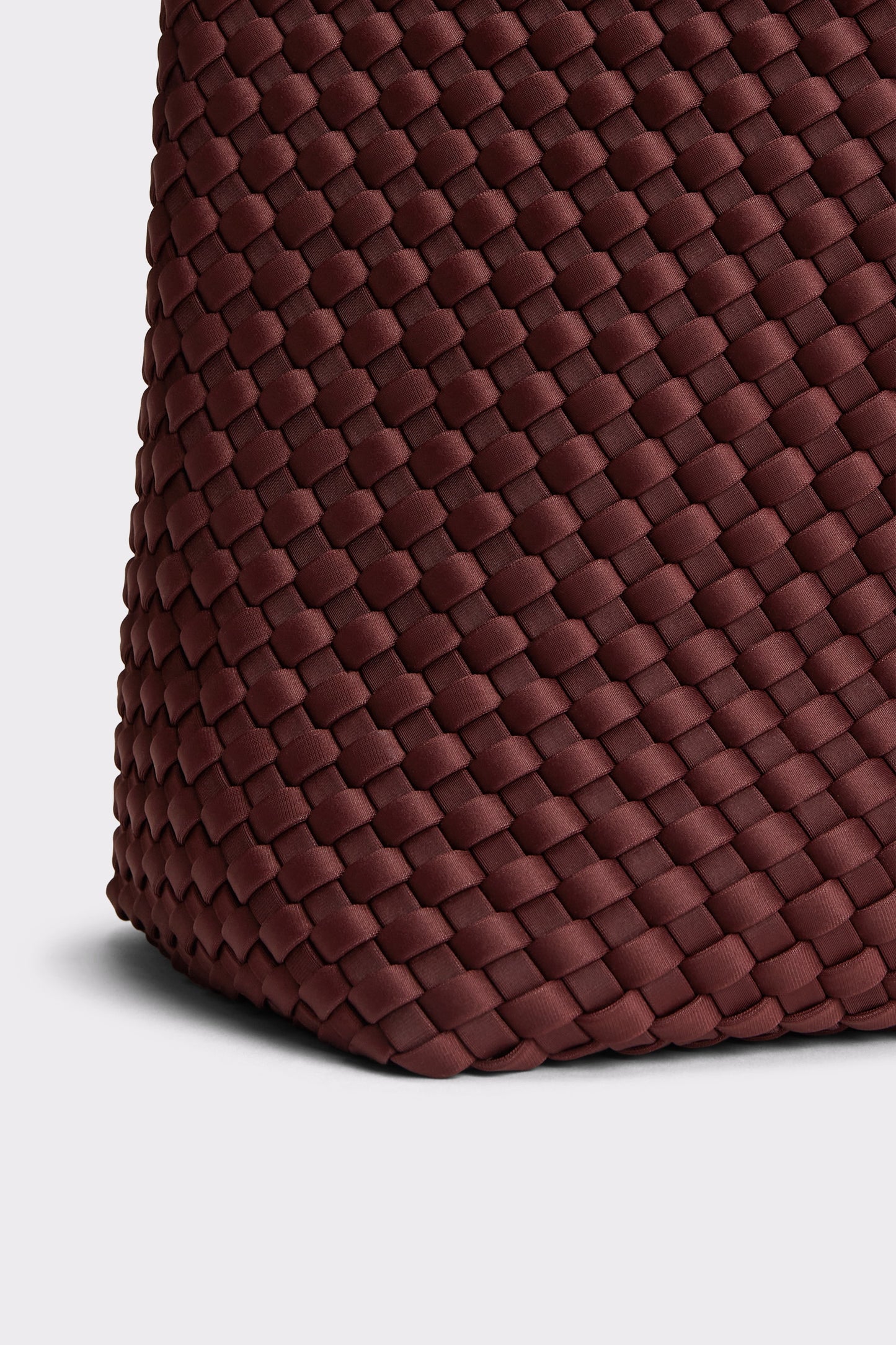 Close-up of a woven leather bag corner in a deep burgundy brown color, showing the detailed basketweave texture against a light gray background.