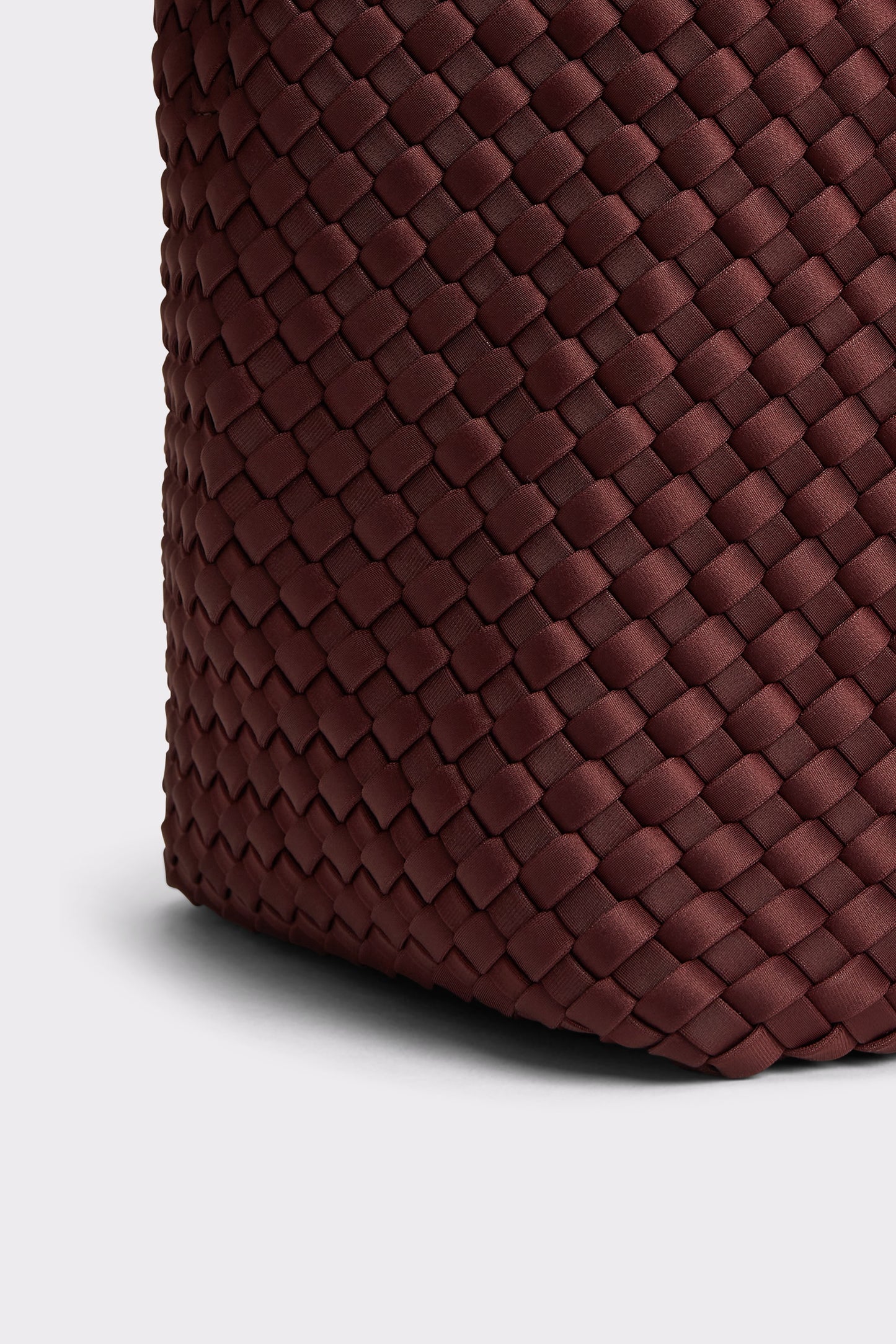 Close-up of a woven leather bag corner in a deep burgundy-brown color, showing the detailed basketweave texture against a light gray background.