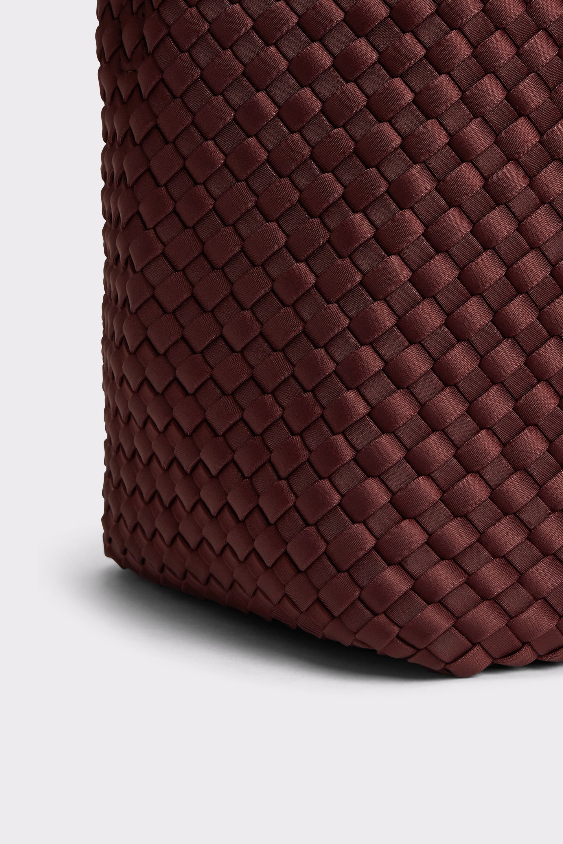 Close-up of a woven leather bag corner in a deep burgundy-brown color, showing the detailed basketweave texture against a light gray background.