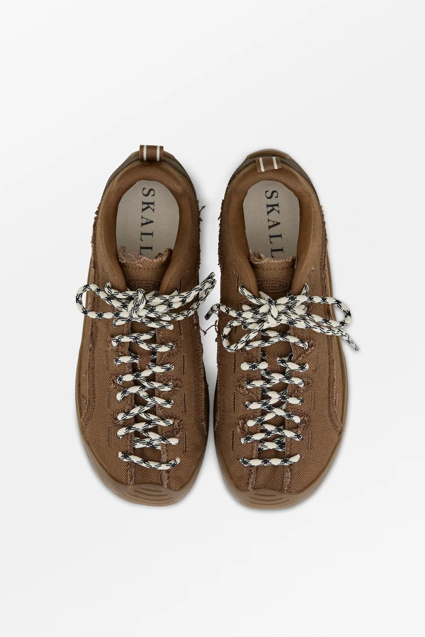 A pair of brown suede low-top sneakers with chunky braided white and black laces, shown from a top-down view on a light gray background. The brand name 'SKULL' is visible on the insole.