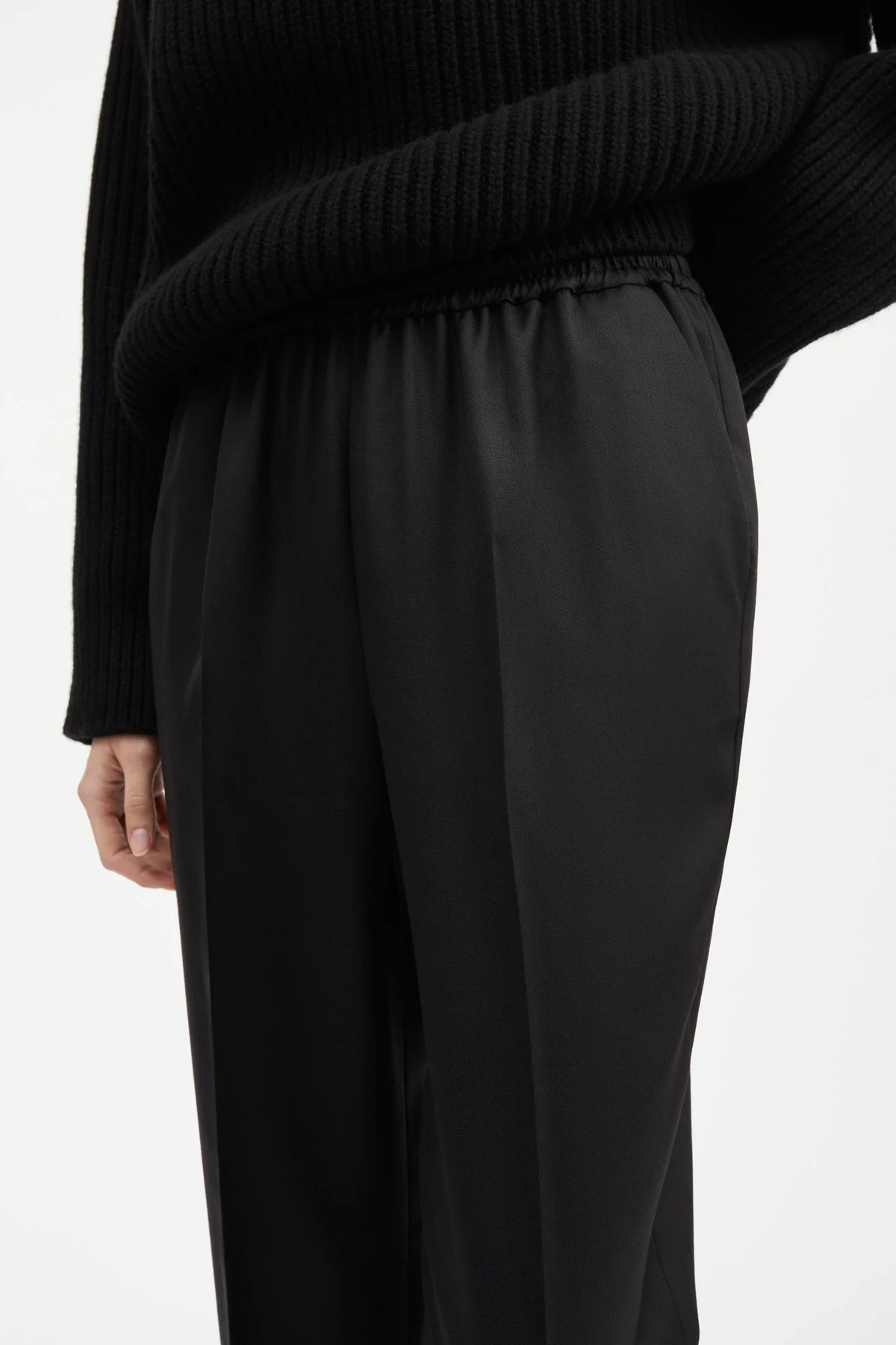 A close-up of a person wearing black wide-leg trousers and a black ribbed knit sweater against a plain light background.