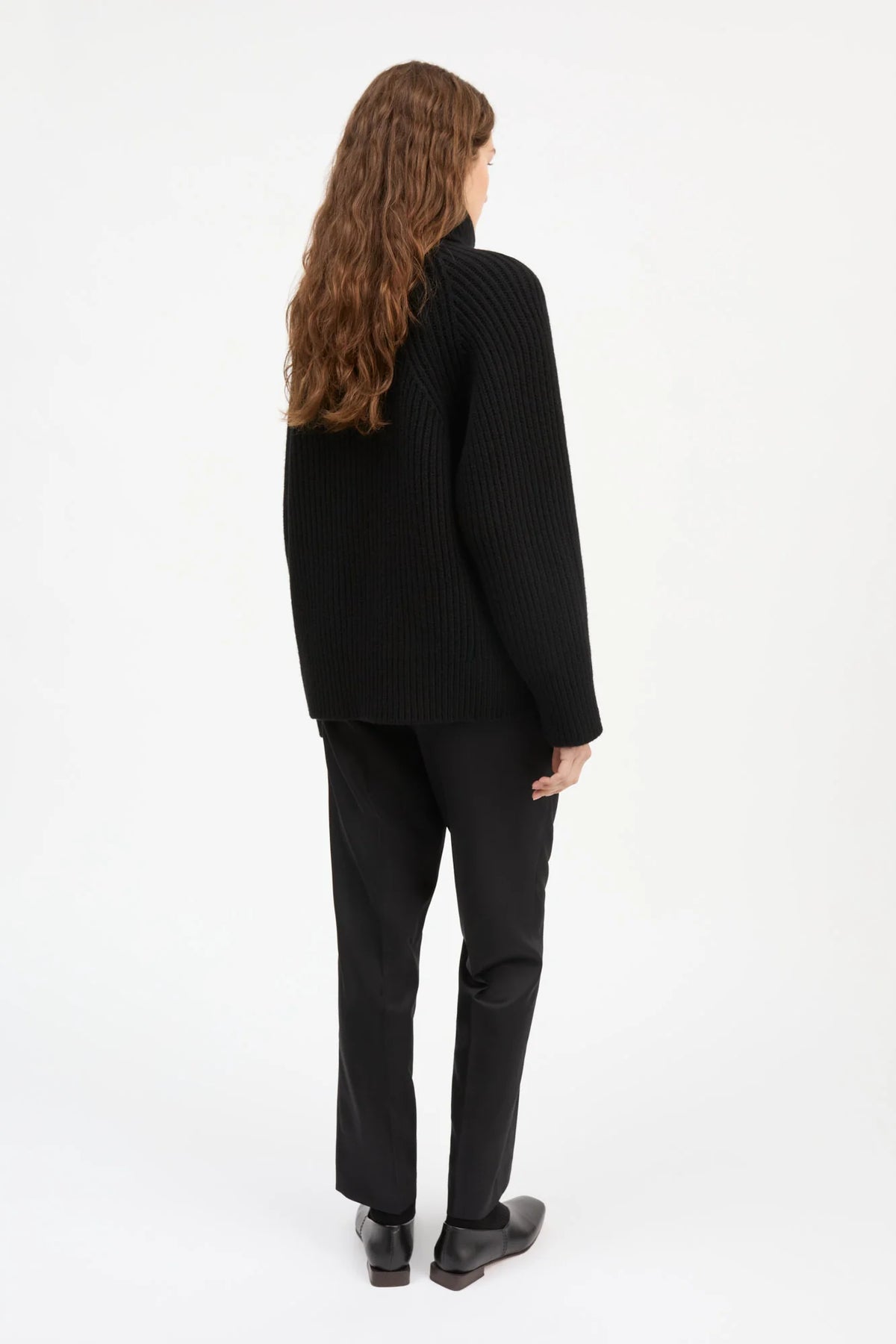 A woman with long wavy brown hair is shown from the back wearing a black ribbed turtleneck sweater and black trousers against a plain white studio background.