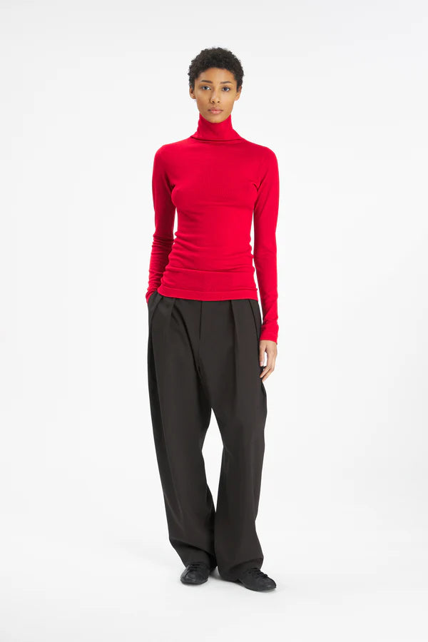A model stands against a plain white studio background wearing a bright red long-sleeve turtleneck top and loose black wide-leg trousers.