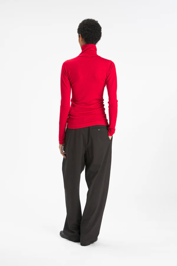 A model shown from the back wearing a bright red long-sleeve turtleneck top and dark charcoal gray wide-leg pants, photographed against a seamless white studio background.