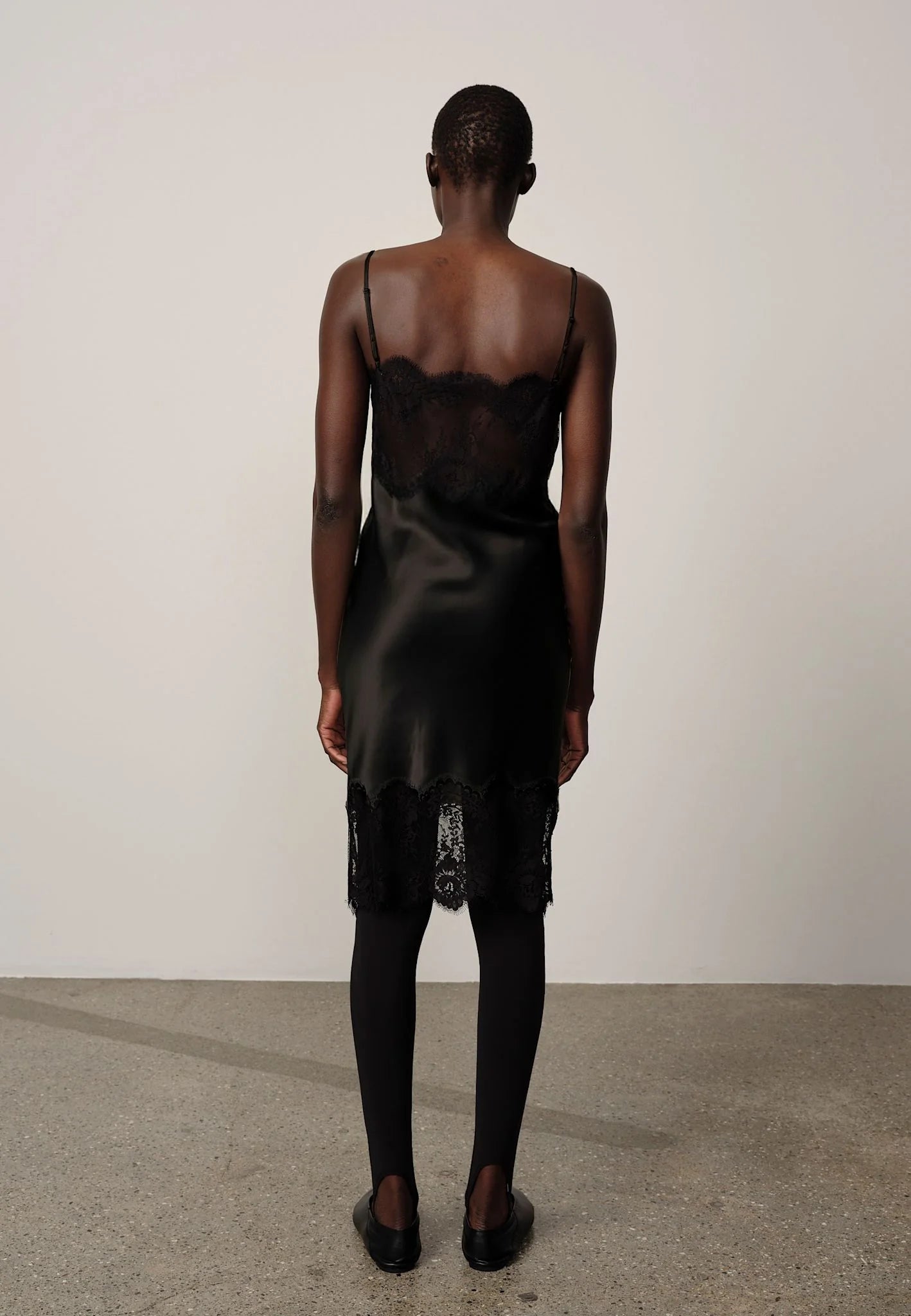 A model viewed from behind wearing a black satin slip dress with a lace bodice and hem, styled with black tights and black shoes against a minimalist studio backdrop.