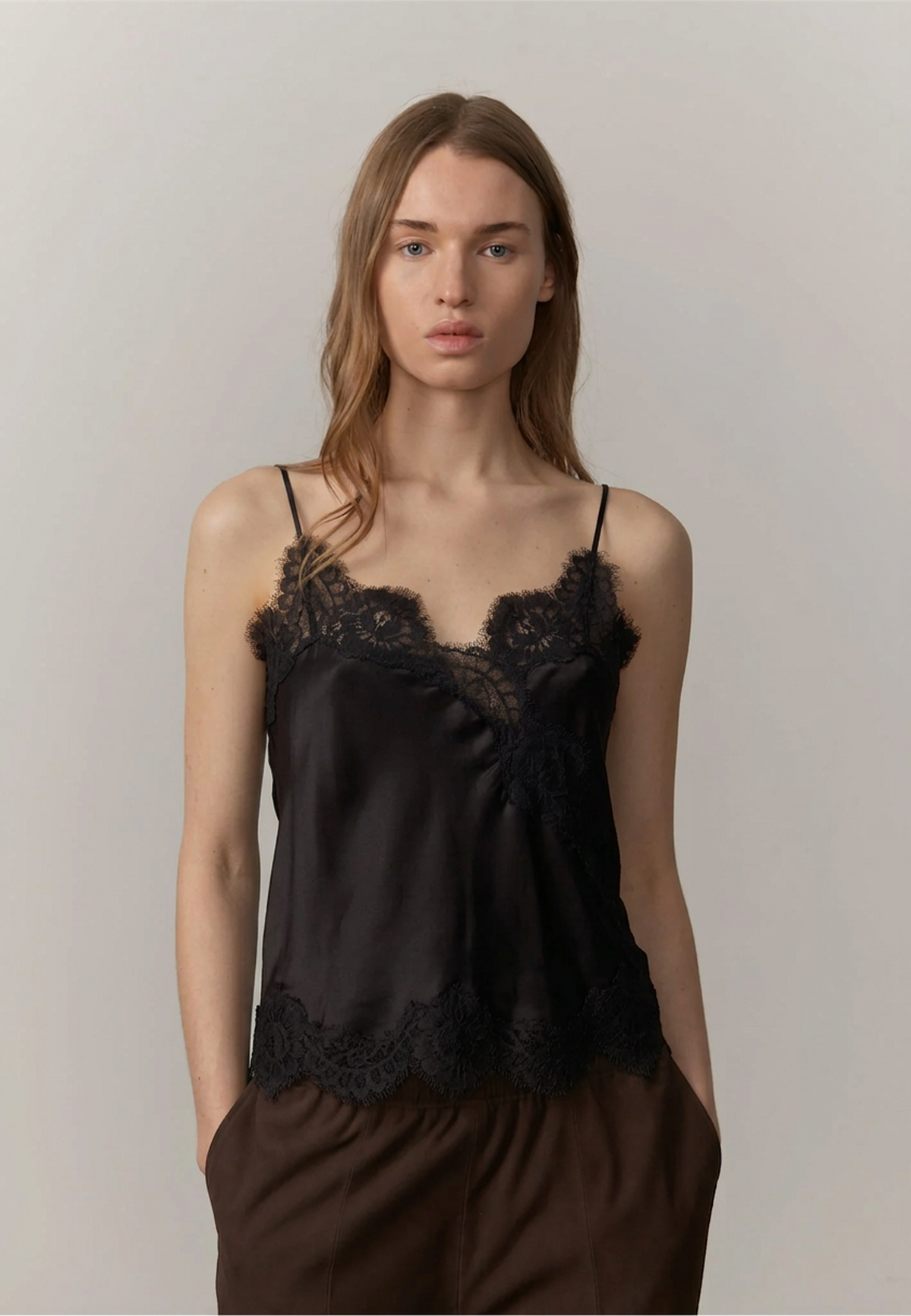 A woman modeling a black satin camisole top with black lace trim along the neckline and hem, paired with brown pants, against a plain light gray studio background.