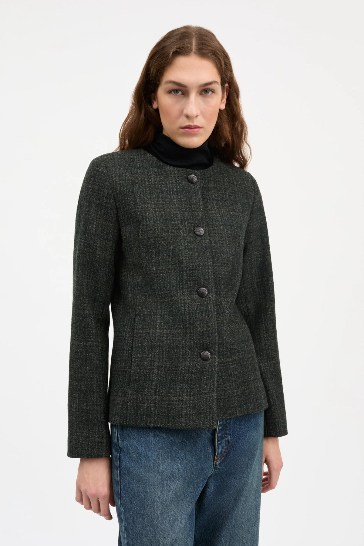 A woman modeling a dark charcoal gray textured tweed jacket with a black mock neck insert, paired with blue jeans, against a plain white studio background.