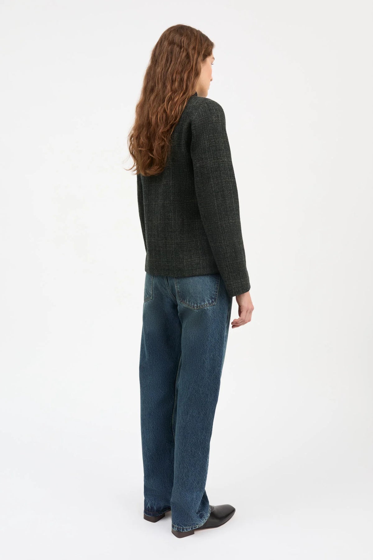 A woman with long wavy brown hair is shown from the back wearing a dark charcoal tweed jacket and blue jeans against a plain white studio background.
