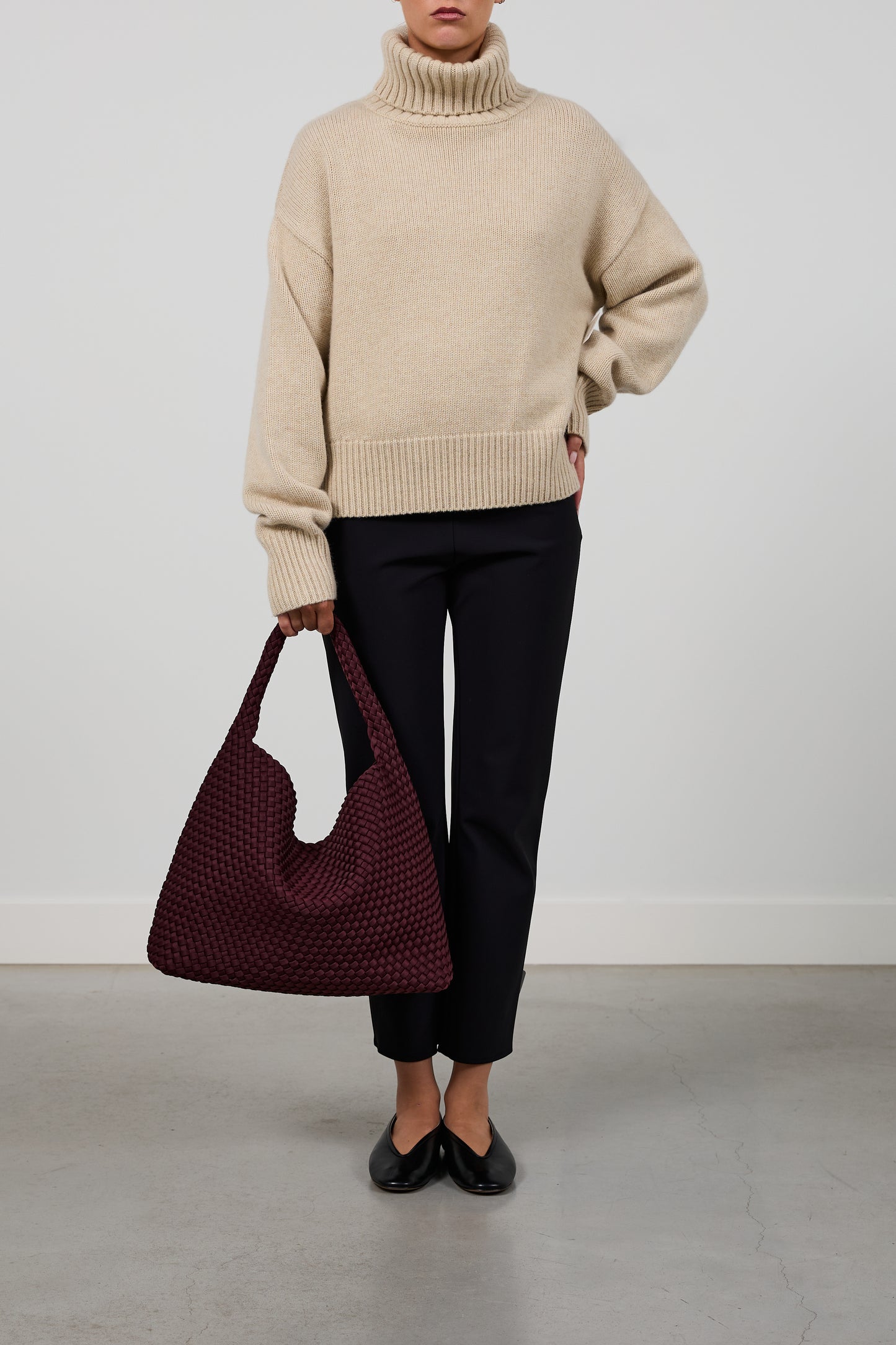 A model wearing a beige ribbed turtleneck sweater, black cropped trousers, and black flats, holding a large burgundy woven shoulder bag against a light gray studio background.