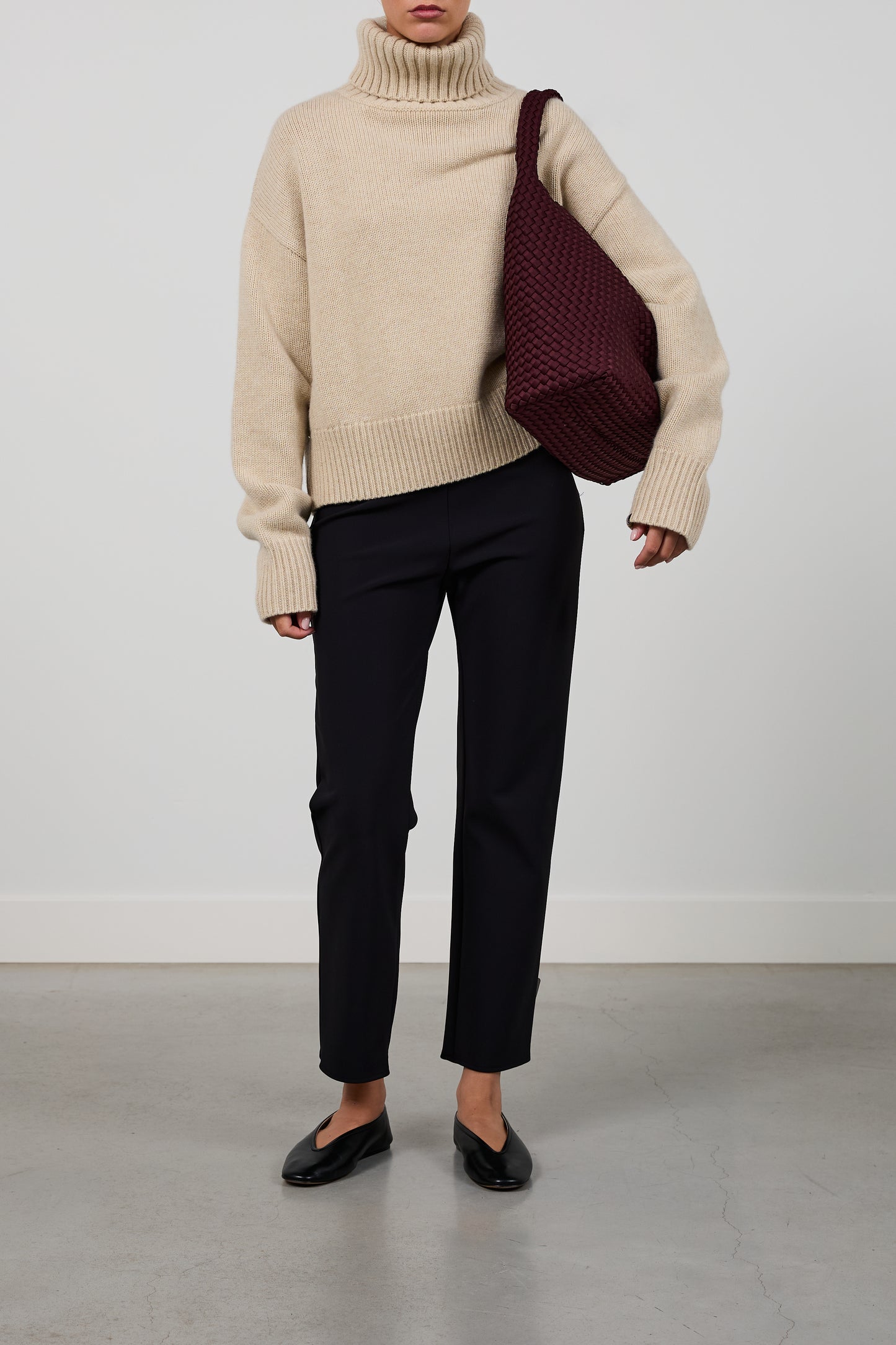 A model wearing a beige turtleneck sweater, black cropped trousers, and black flats, carrying a large burgundy woven shoulder bag against a plain light gray studio background.