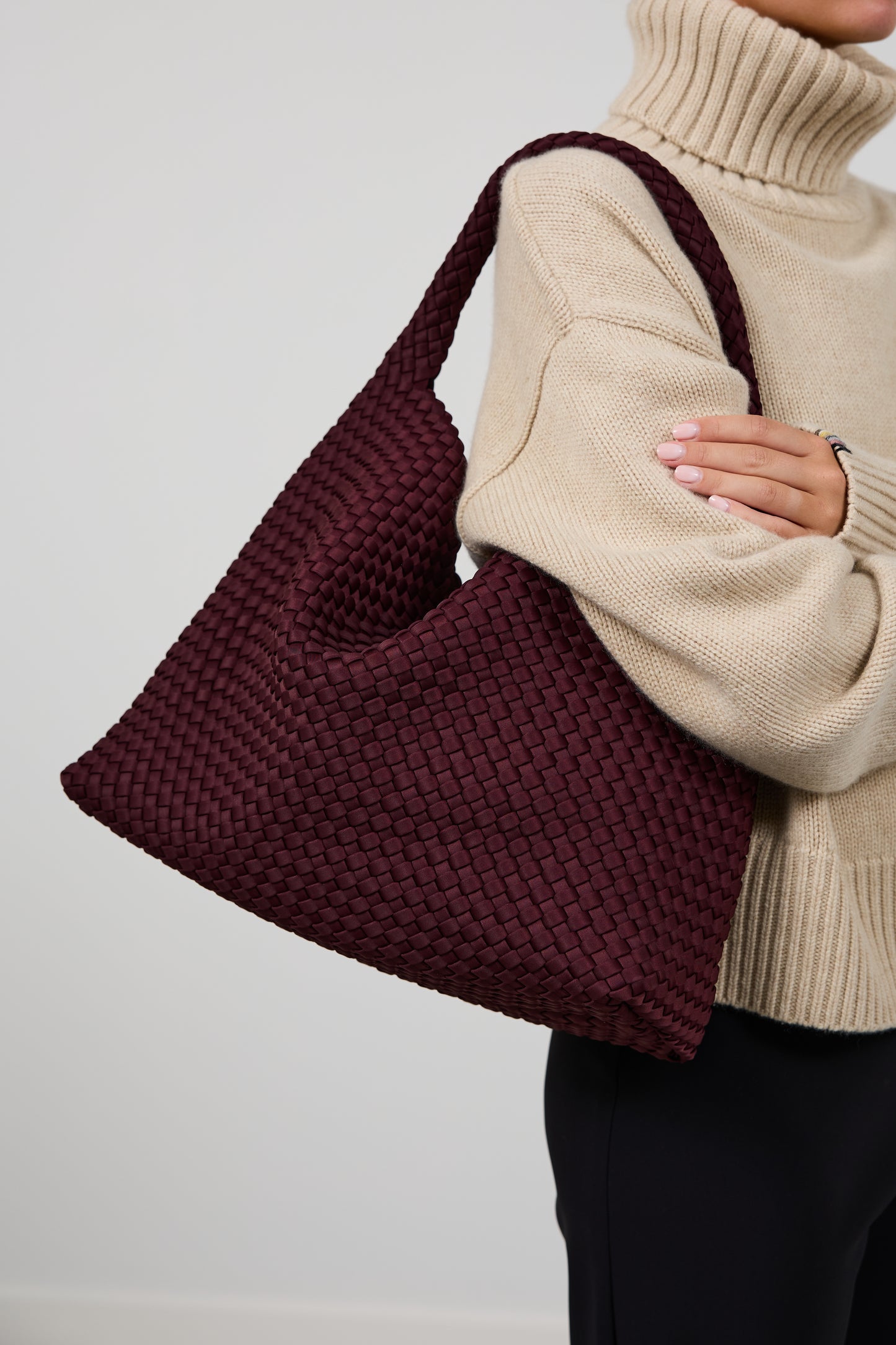 A person wearing a beige ribbed turtleneck sweater and black pants carries a large woven shoulder bag in deep burgundy. The bag has a wide, slouchy shape with a matching shoulder strap and a textured, basketweave-like knit pattern.