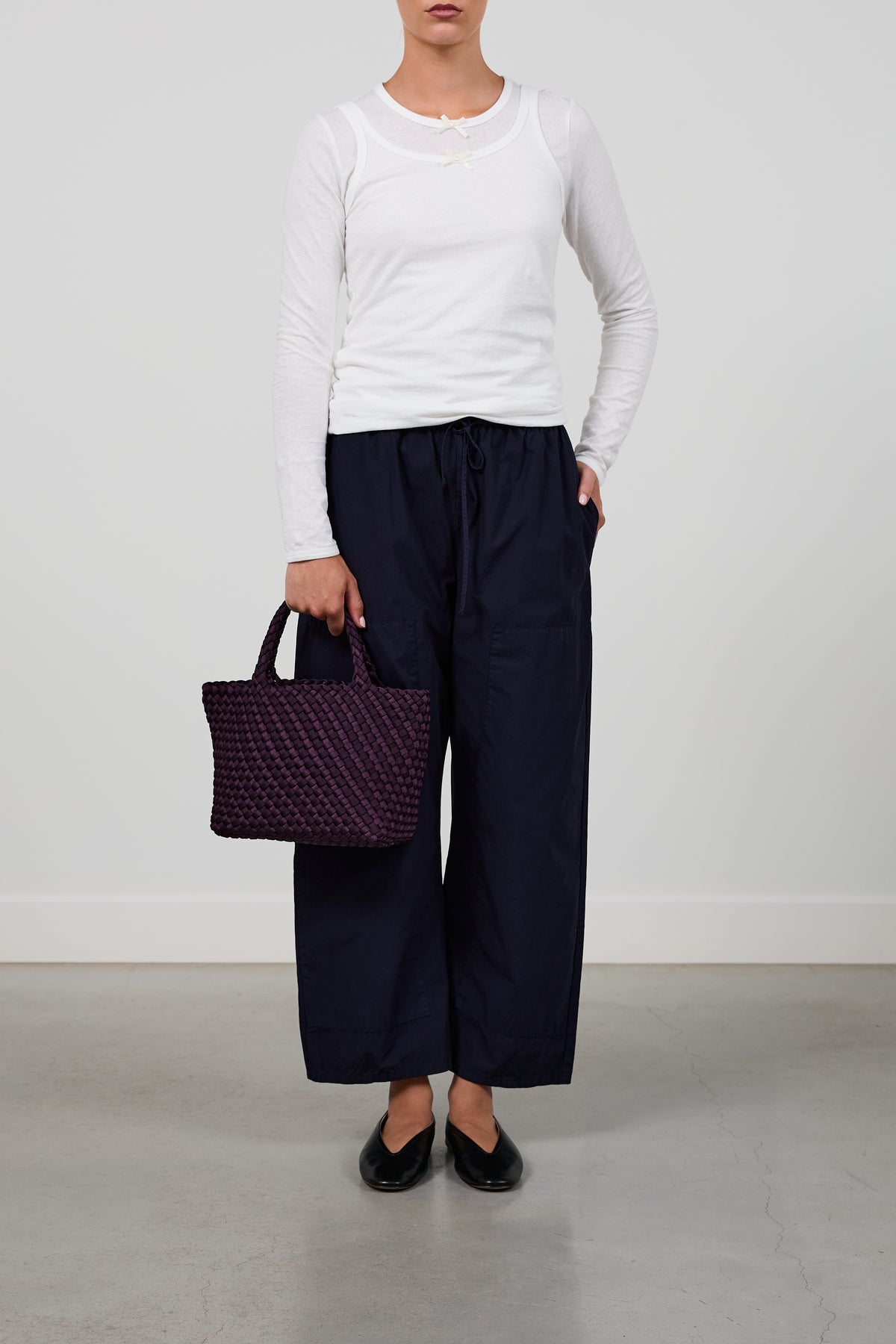 A model wearing a white long-sleeve top, dark navy wide-leg trousers, and black flats, holding a purple woven tote bag against a plain light gray studio background.