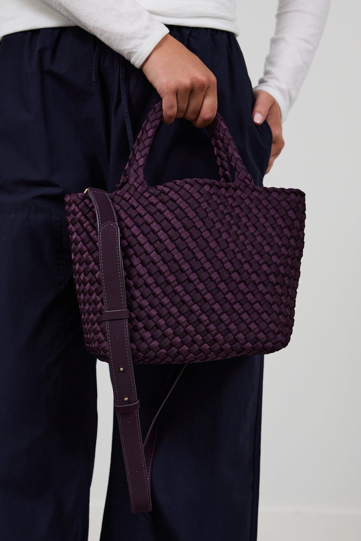 A person wearing dark navy pants and a white long-sleeve top holds a woven dark purple tote bag with top handles and a matching shoulder strap against a plain light background.