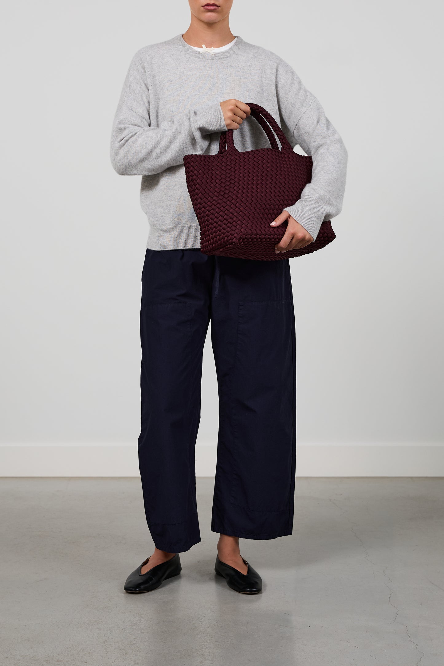 A model wearing a light gray crewneck sweater, dark navy wide-leg trousers, and black flats, holding a burgundy woven tote bag against a plain light gray studio background.