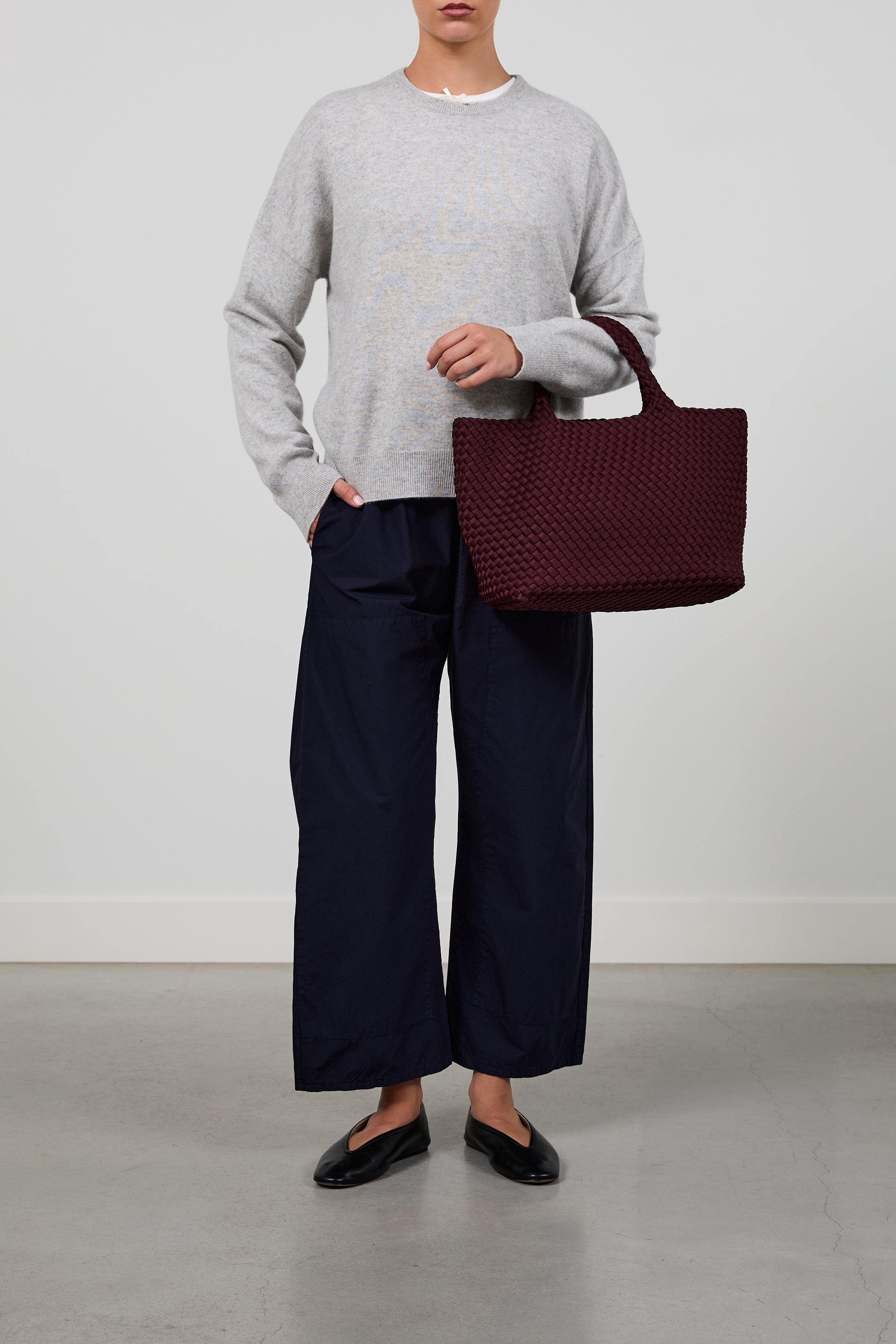 A model wearing a light gray crewneck sweater, dark navy wide-leg trousers, and black flats, holding a large burgundy woven tote bag against a plain studio background.