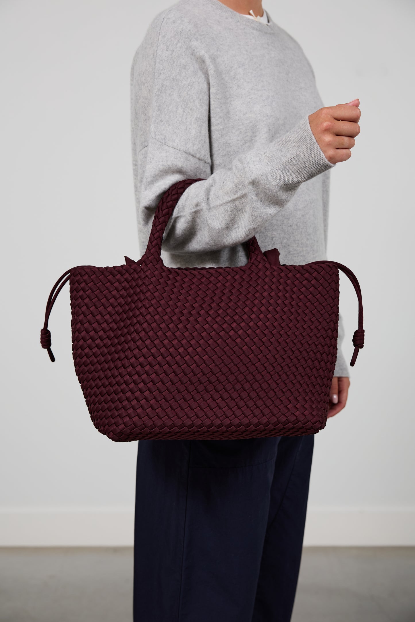 A person wearing a light gray sweater and navy pants carries a large woven tote bag in deep burgundy. The person is cropped from the neck down, standing against a plain light gray wall.