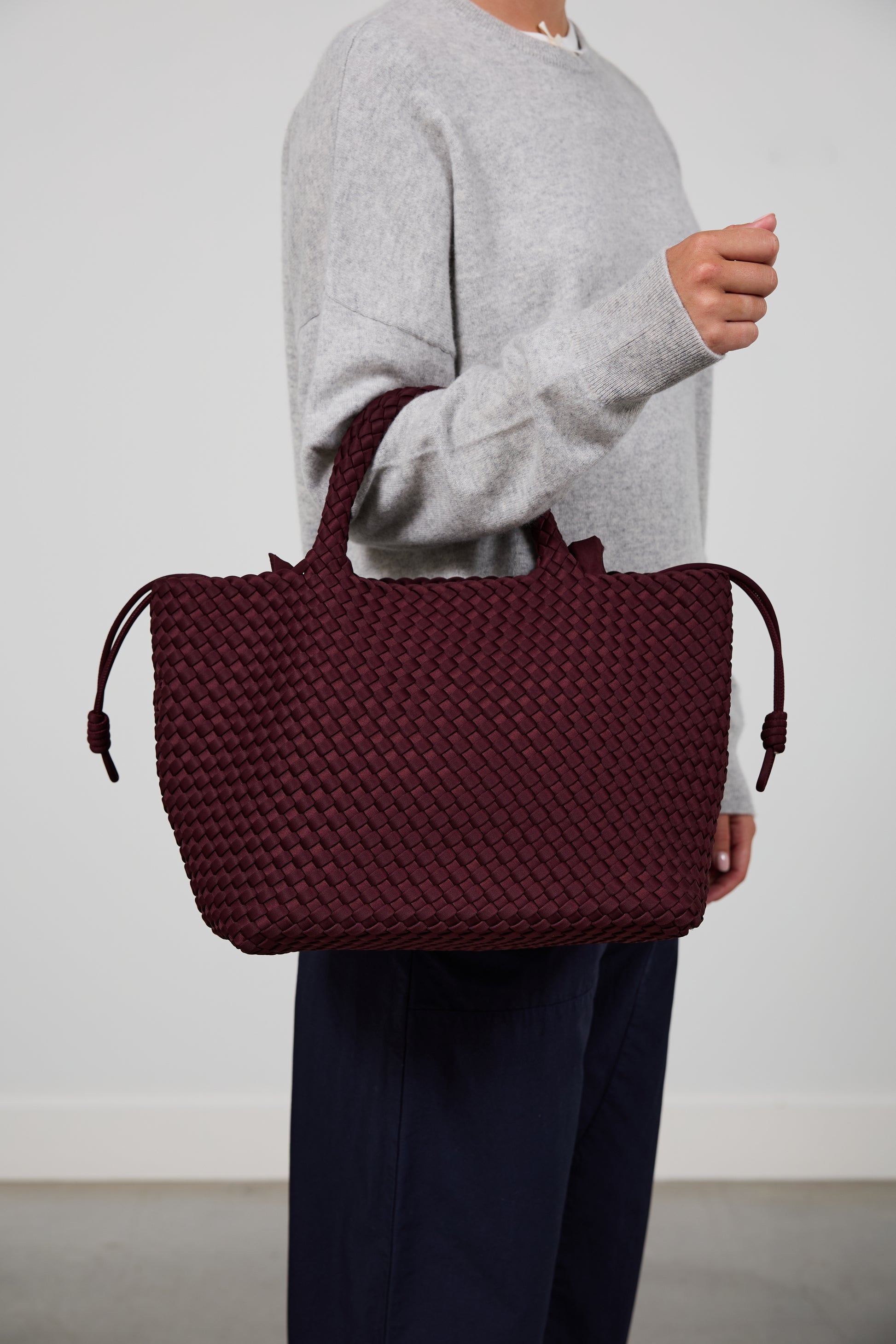 A person wearing a light gray sweater and navy pants carries a large woven tote bag in deep burgundy. The person is cropped from the neck down, standing against a plain light gray wall.