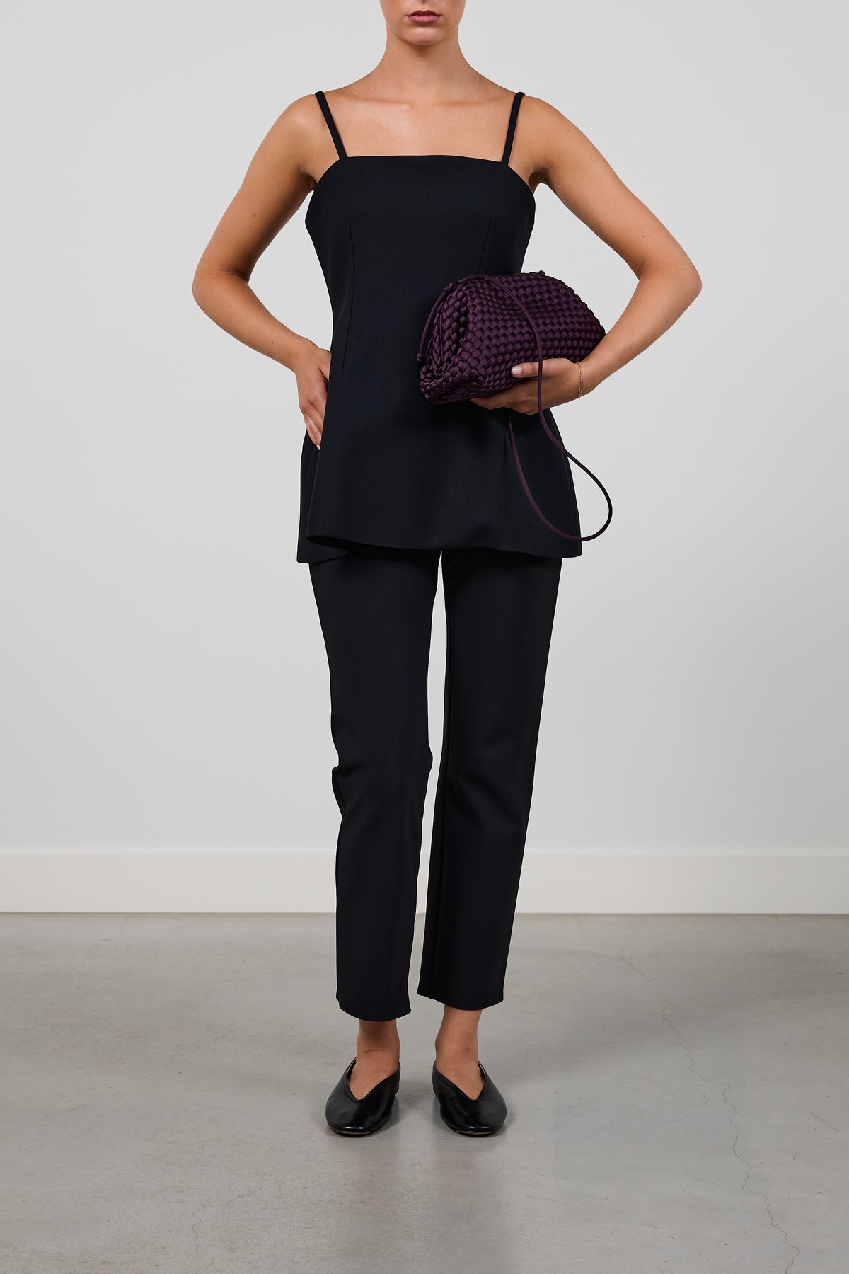 A model wearing a black camisole top and matching black trousers, holding a dark purple woven clutch bag, set against a plain light gray studio background.