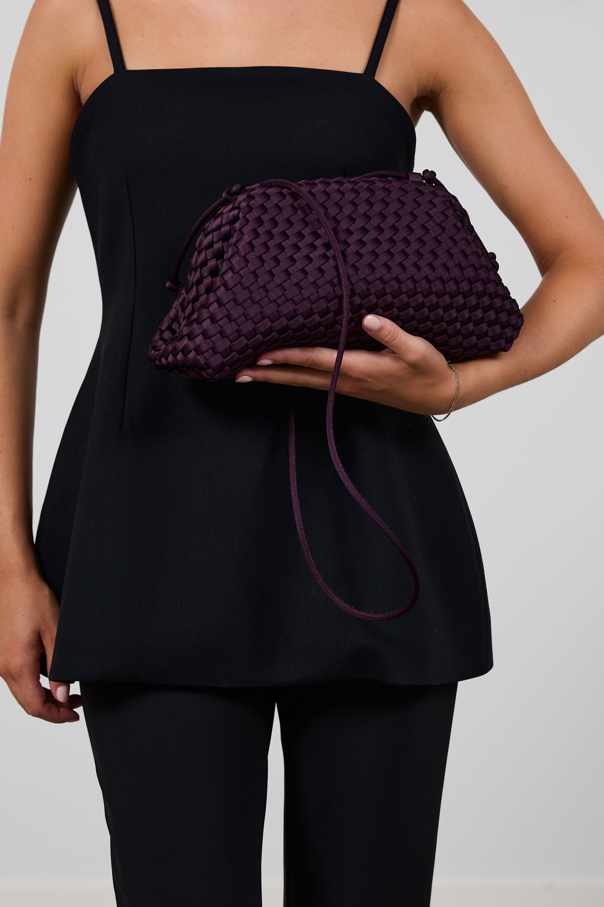 A woman wearing a black sleeveless top and black leggings holds a dark purple woven clutch bag with a thin strap against a light gray background.