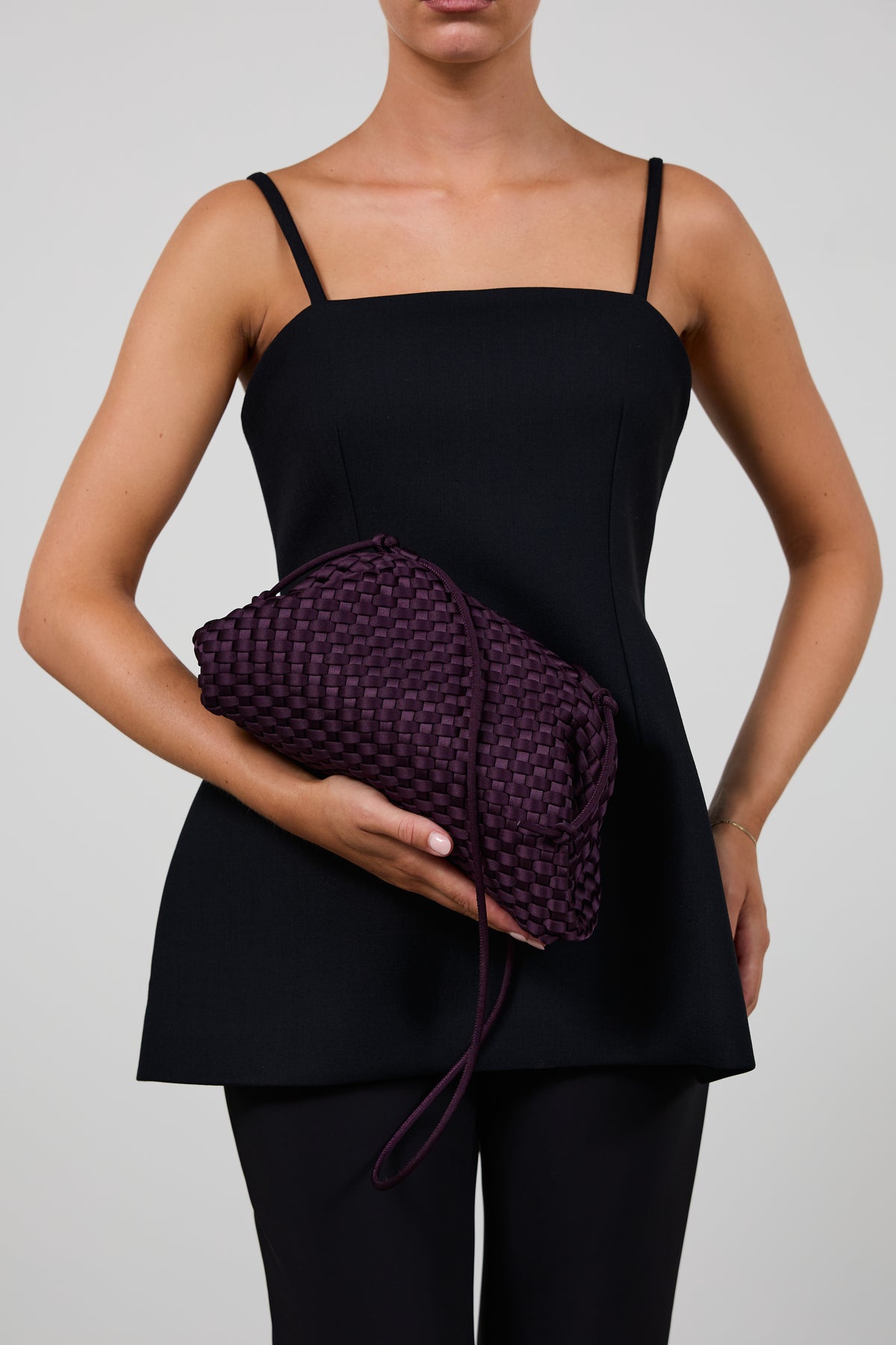 A model wearing a black camisole top and black pants holds a dark purple woven clutch bag against a light gray studio background.