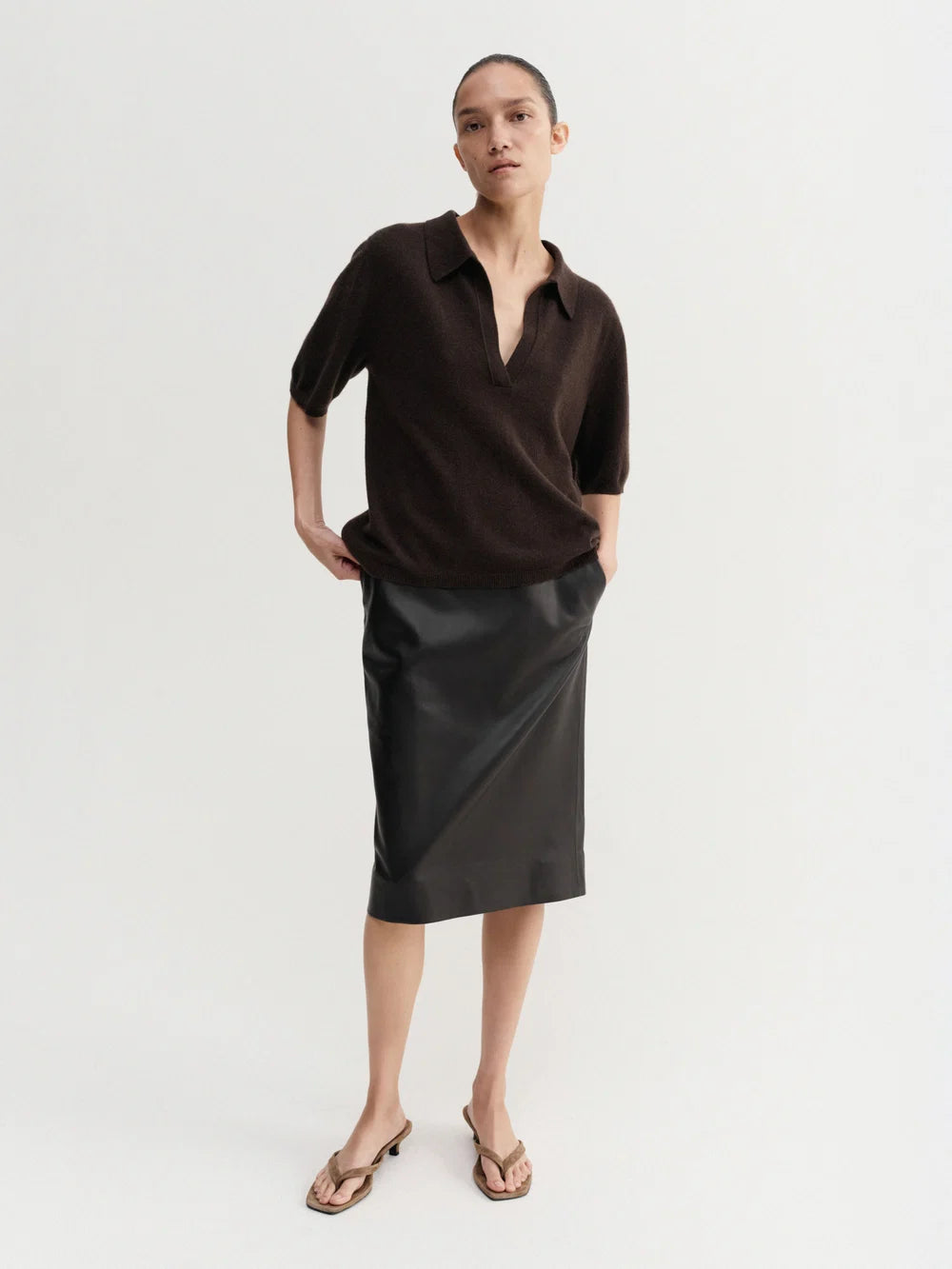 A model stands against a plain light gray studio background wearing a dark brown short-sleeve polo-style top and a black midi skirt, with tan heeled sandals.