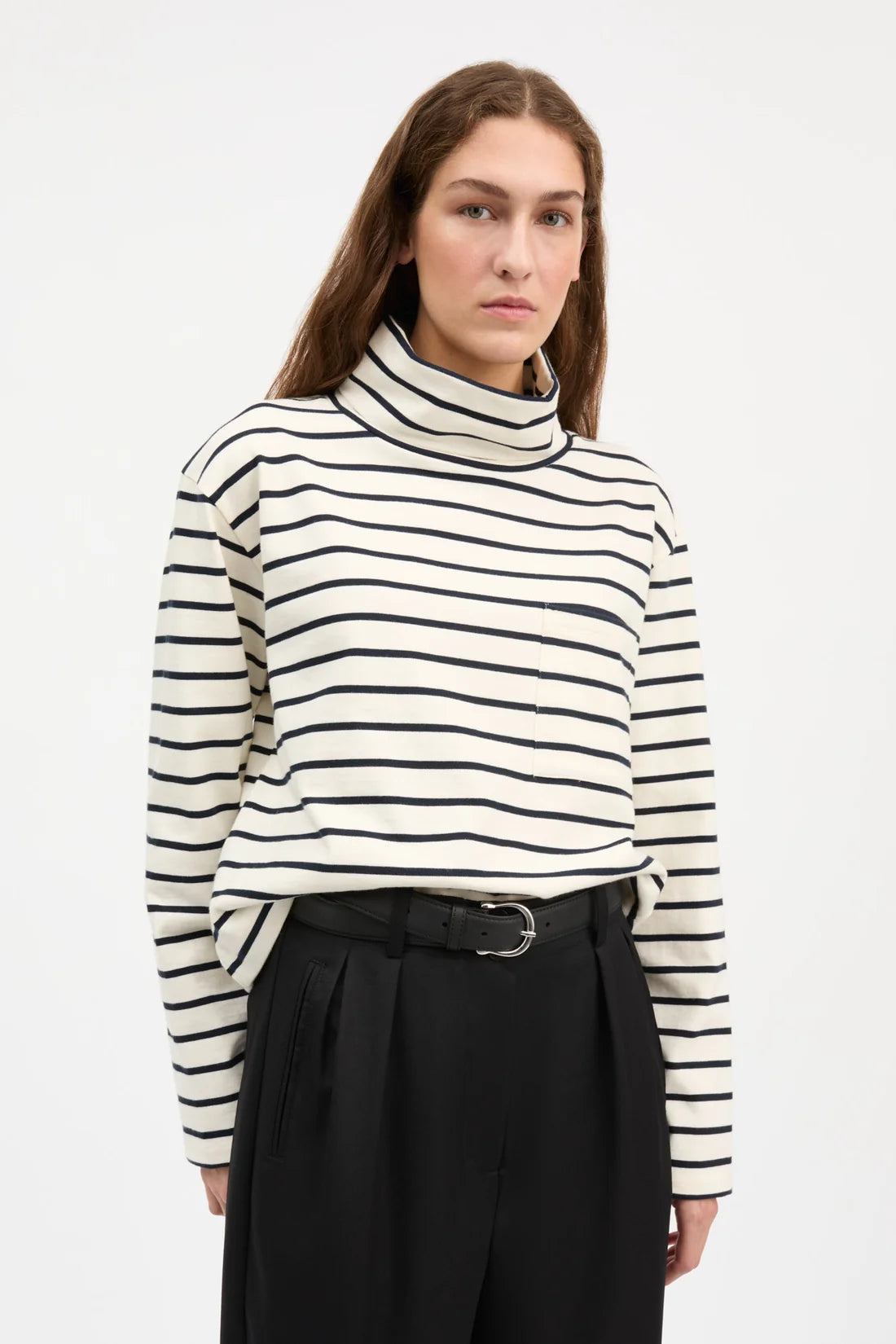A woman wearing a cream and black horizontally striped mock neck sweater, paired with black trousers and a black belt, against a plain white studio background.