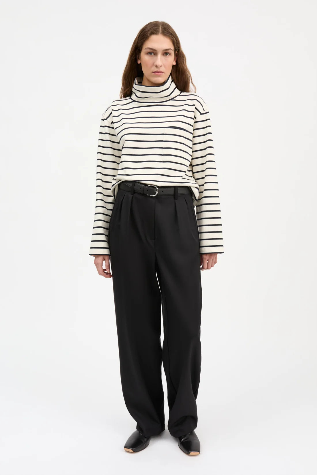 A woman modeling a black and white horizontally striped long-sleeve turtleneck sweater paired with black high-waisted trousers and black shoes, shot against a plain white studio background.