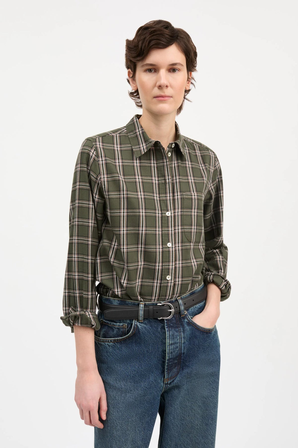 A model stands against a plain white studio background wearing a long-sleeve green and brown plaid button-down shirt, blue jeans, a dark belt