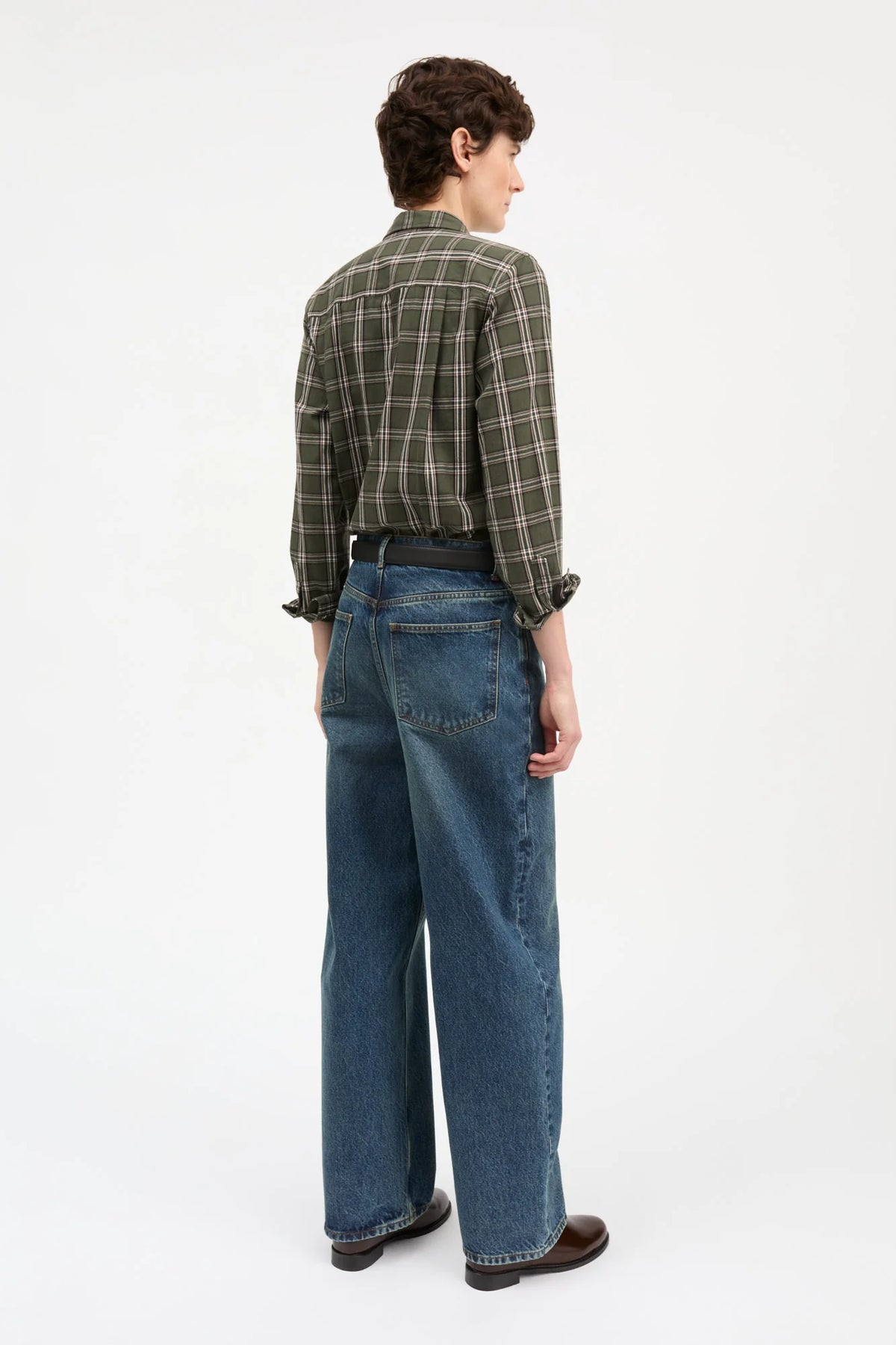 A model stands against a plain white studio background wearing a long-sleeve green and brown plaid button-down shirt, blue jeans, a dark belt, and brown leather shoes.