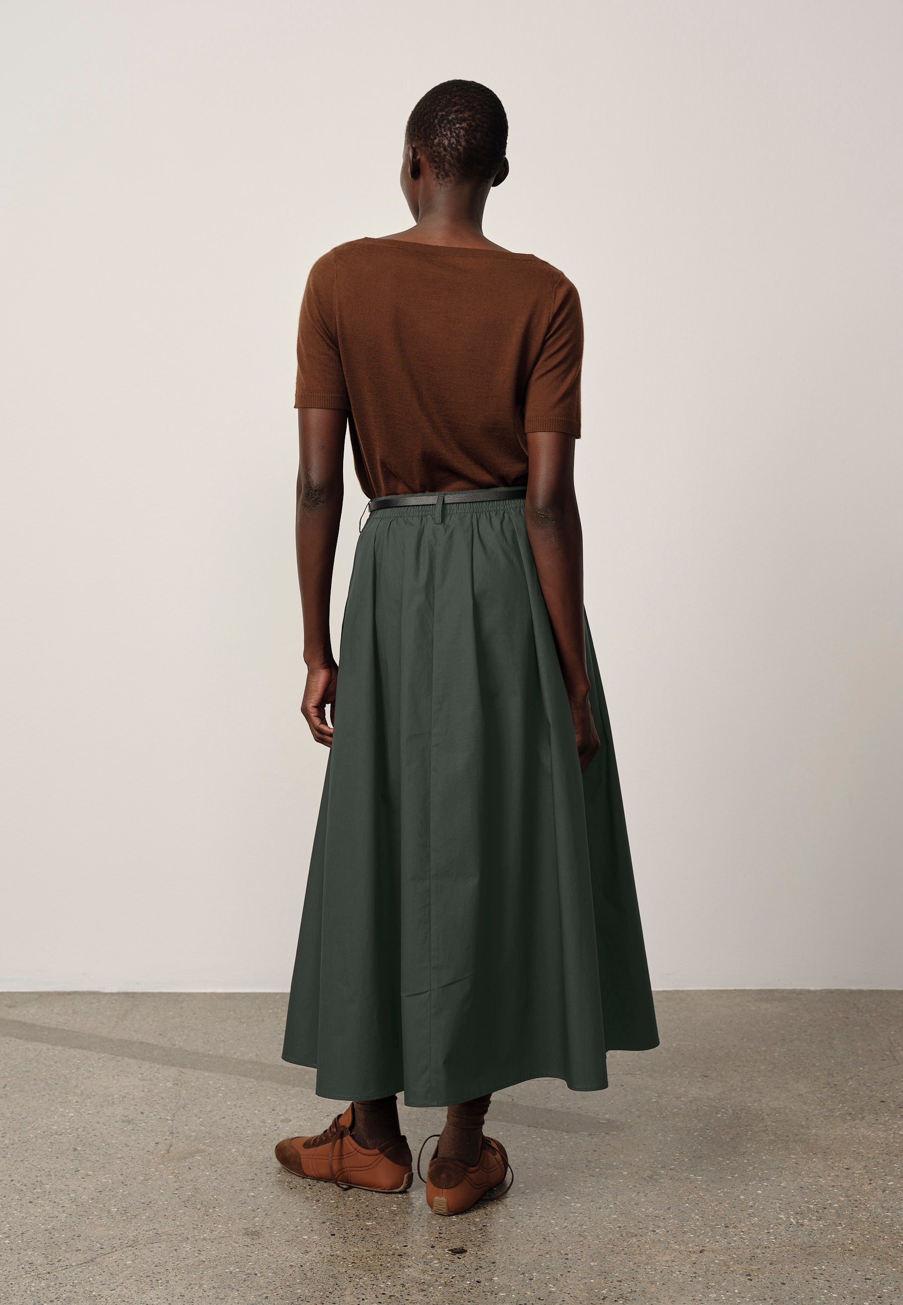 Person wearing a brown top and green skirt standing against a white wall.