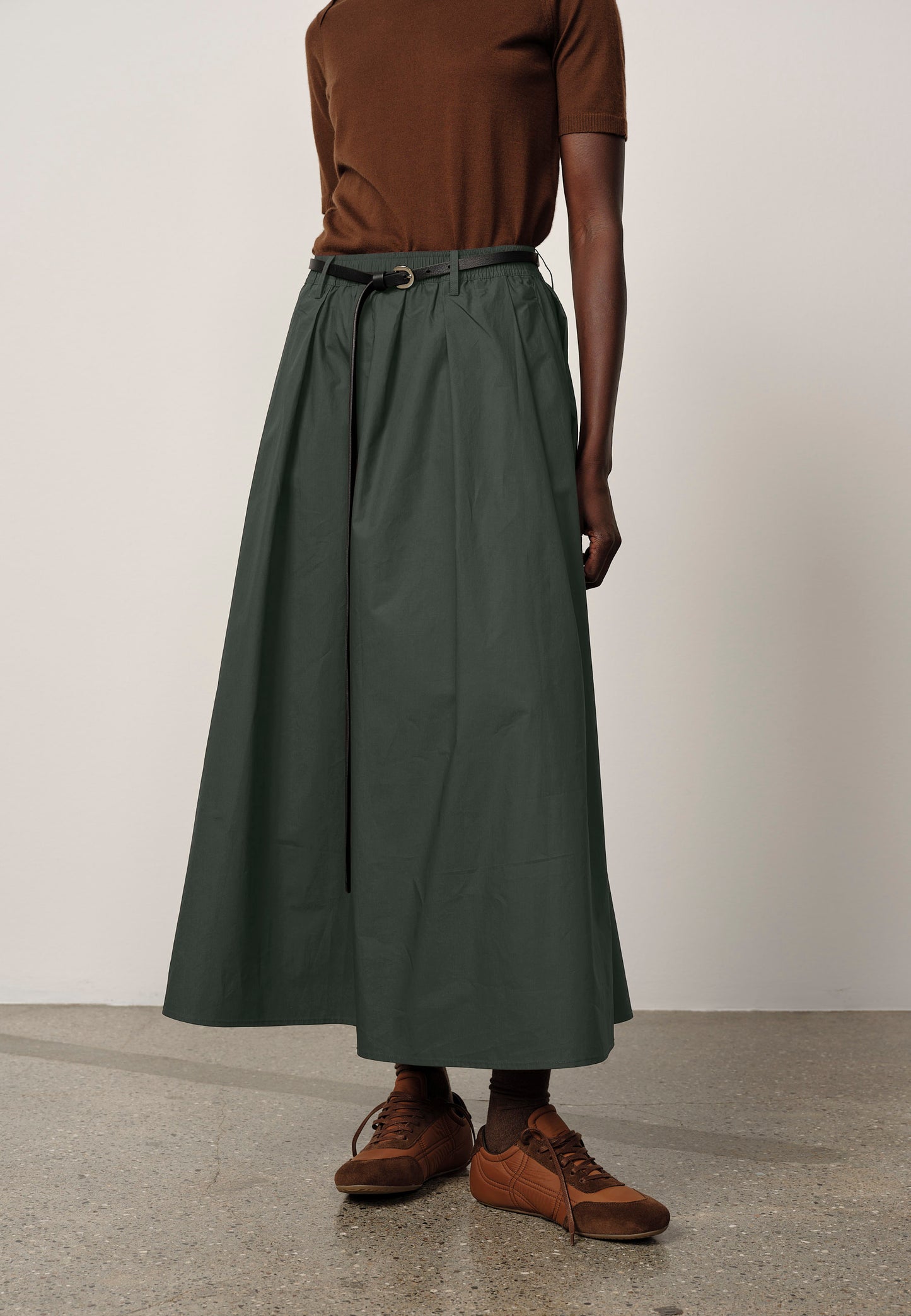 Person wearing a long green skirt with a brown top and shoes on a plain background