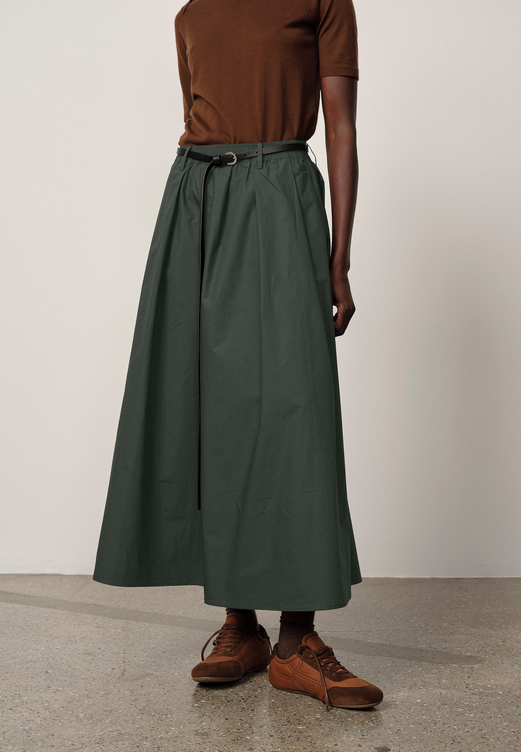 Person wearing a long green skirt with a brown top and shoes on a plain background