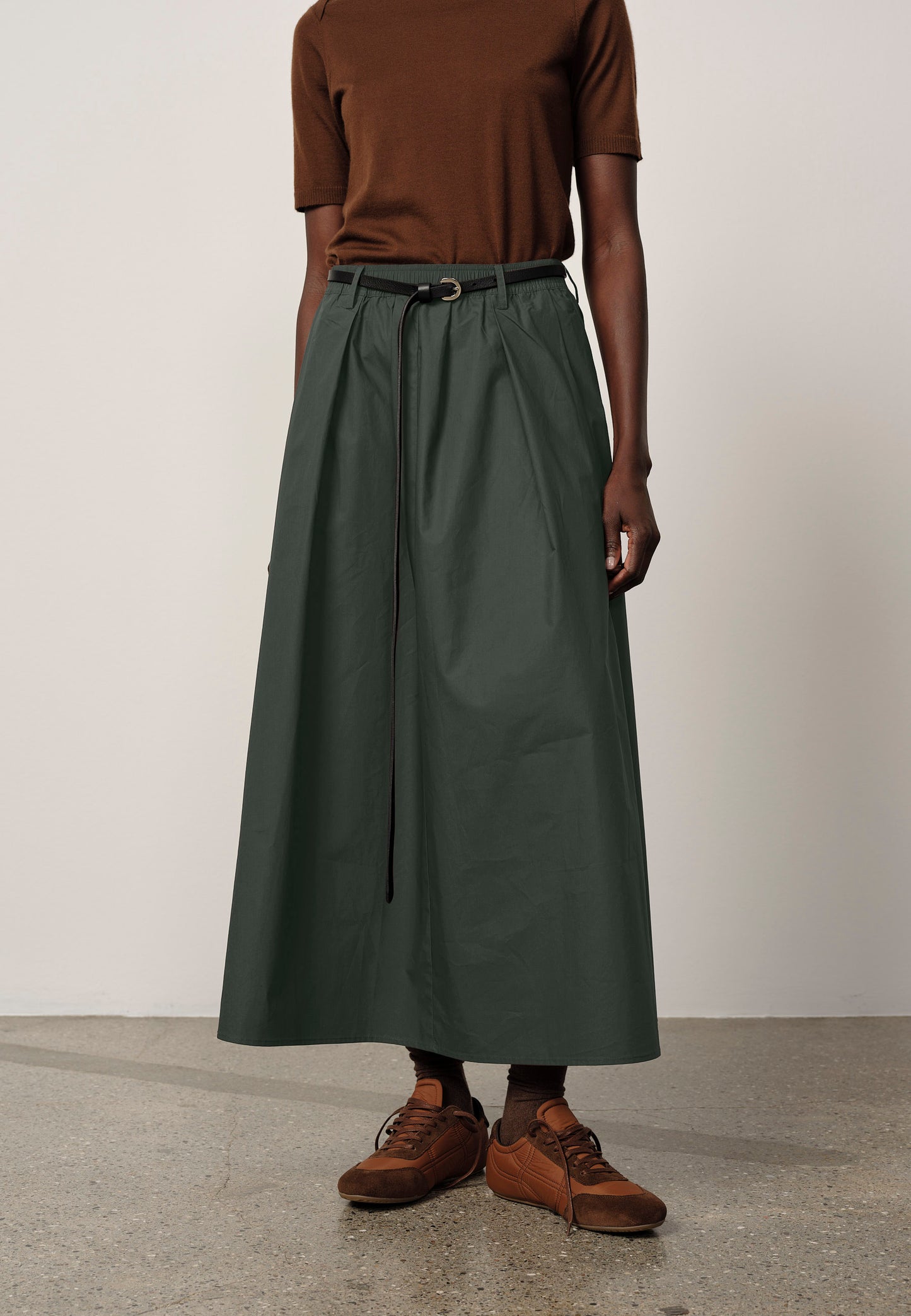 Person wearing a brown top and green skirt with brown shoes on a plain background