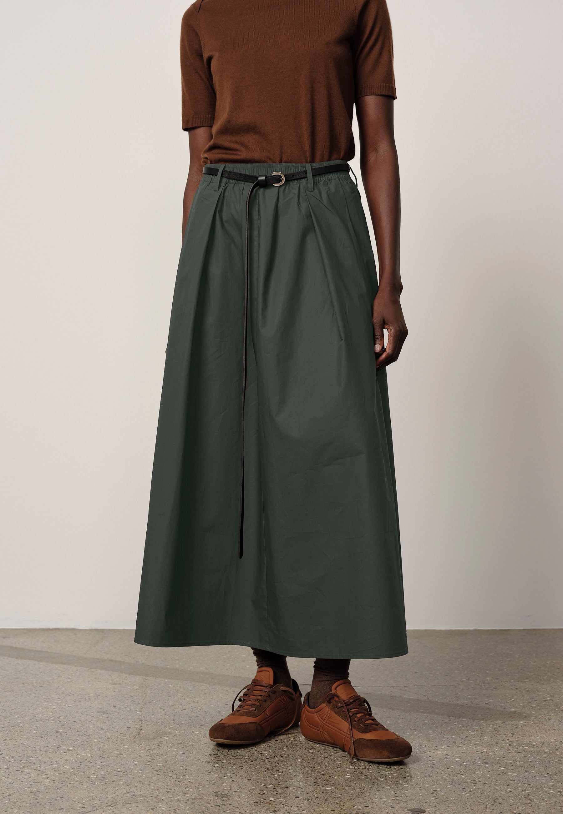Person wearing a brown top and green skirt with brown shoes on a plain background