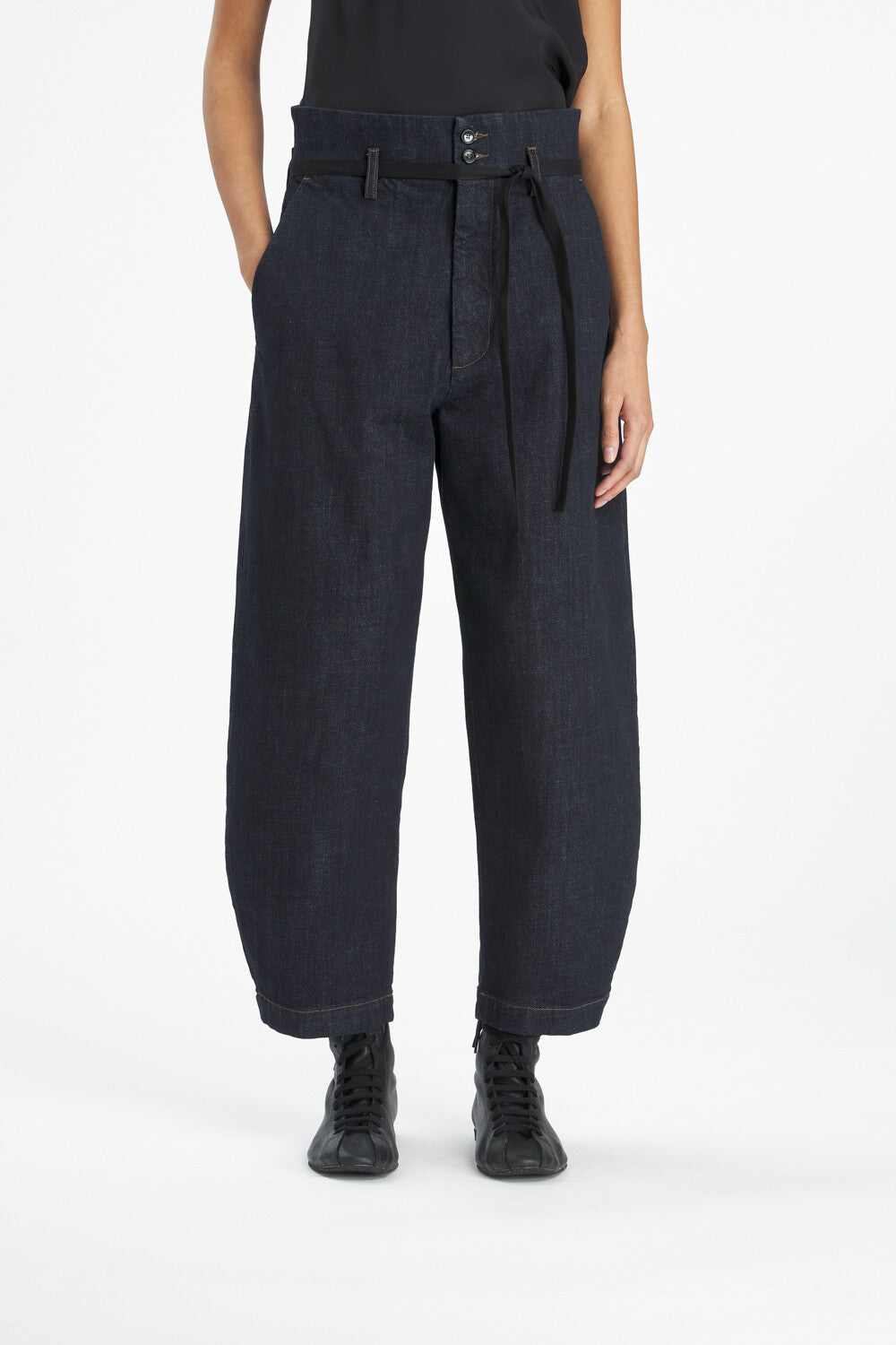 A model from the waist down wearing dark indigo wide-leg denim pants with a matching tie belt, paired with black sneakers against a plain white studio background.