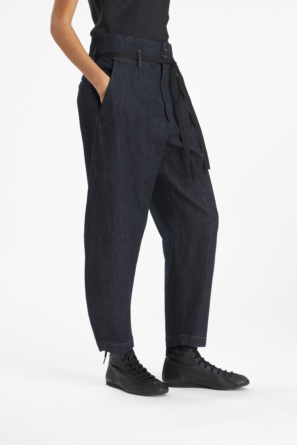 A person wearing dark indigo denim paperbag pants with a matching belt, paired with black sneakers against a clean white studio background.