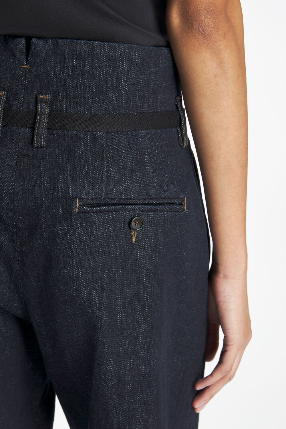 Close-up of a person wearing dark indigo denim trousers with a black belt and a buttoned welt pocket on the back.