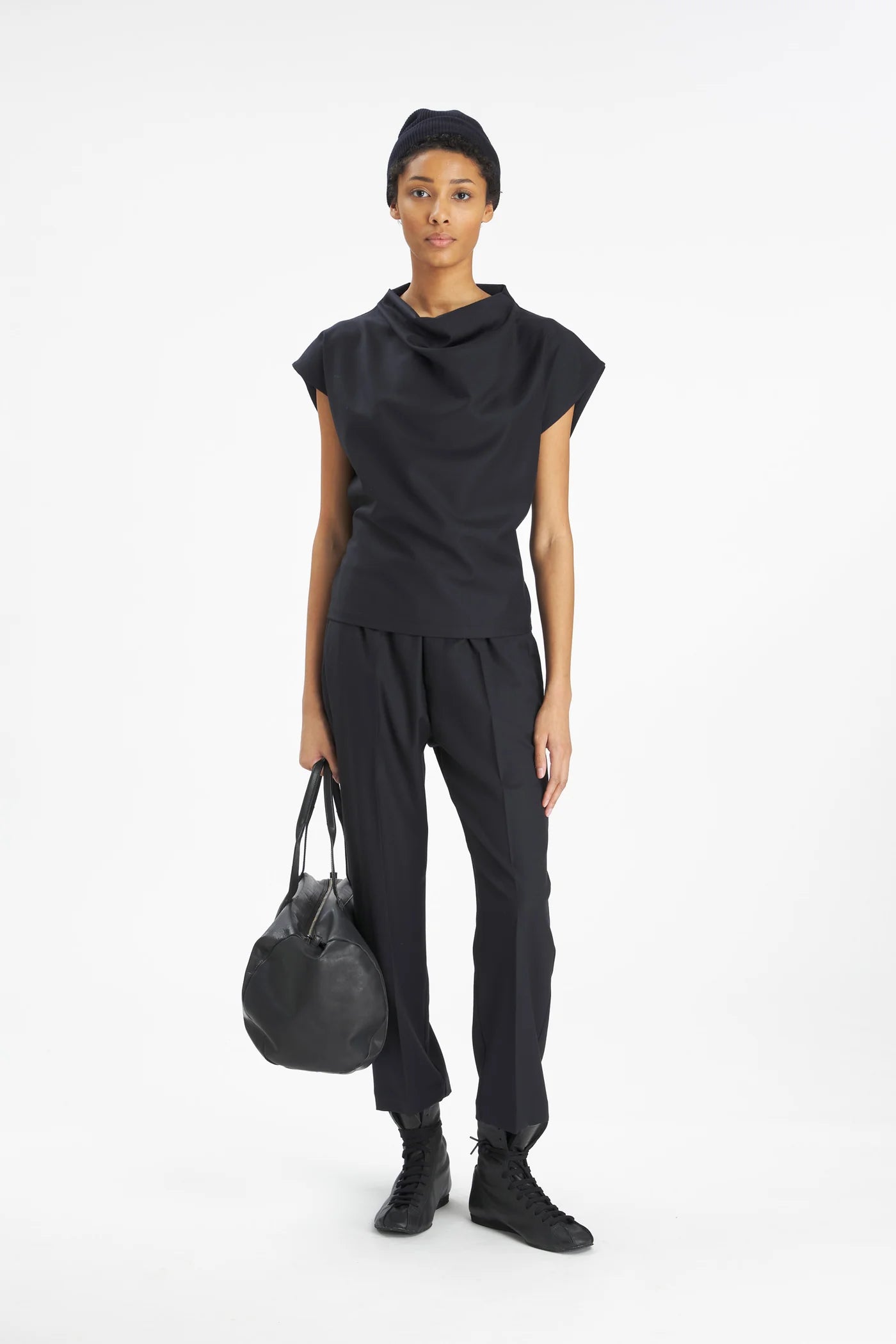 A model wearing a black cowl-neck short-sleeve top, matching black trousers, black sneakers, and a black beanie, holding a black leather duffel bag against a plain white studio background.