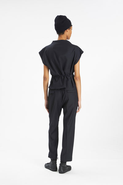 A model shown from the back wearing a black short-sleeve top with a gathered waist, black trousers, and a black beanie against a plain white studio background.