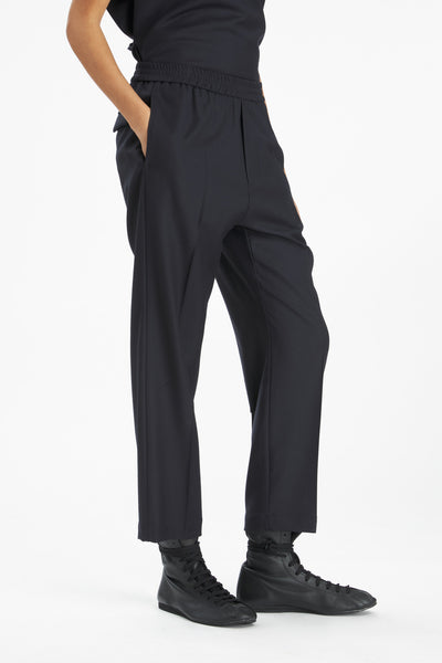 A person wearing black wide-leg cropped trousers with an elastic waistband, paired with black shoes, shown against a plain light gray studio background.
