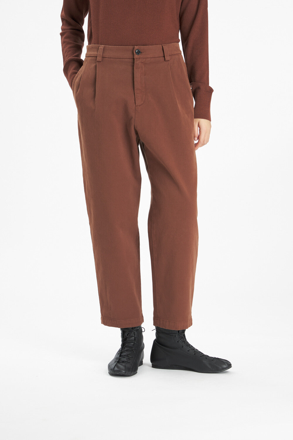 A person wearing brown pleated trousers, a matching brown long-sleeve top, and black lace-up boots, photographed against a clean white studio background.
