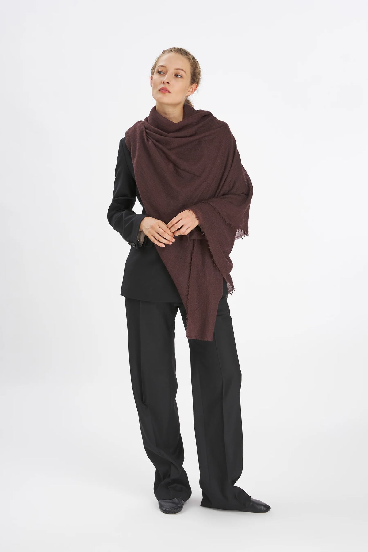 A model wearing a dark brown shawl draped over her shoulders, paired with a black long-sleeve top and black wide-leg trousers, standing against a plain white studio background.