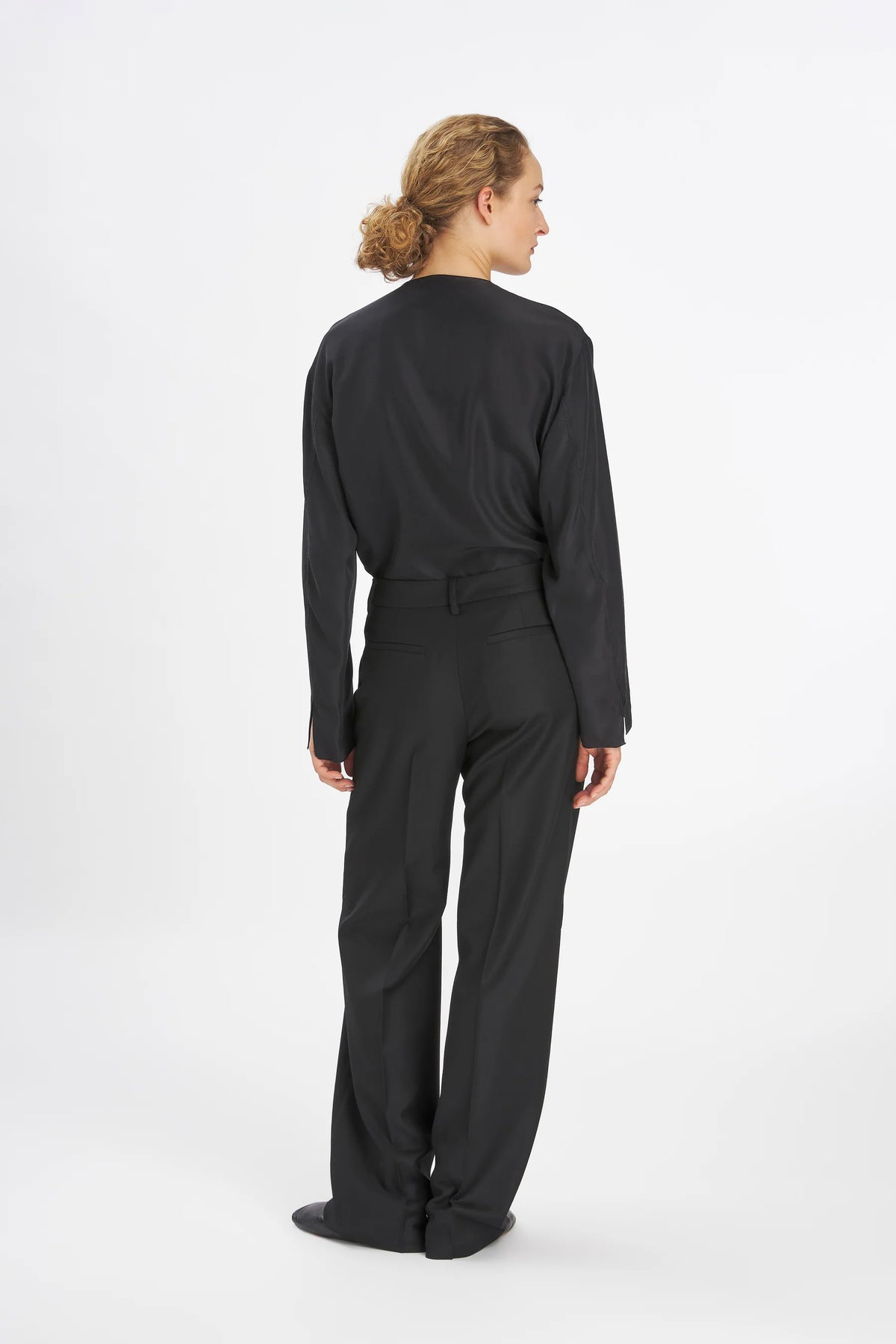 A model shown from the back wearing a black long-sleeve top and matching black wide-leg trousers against a plain white studio background.