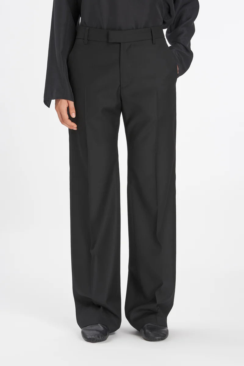 A person wearing black wide-leg trousers with a matching black belt, paired with a black long-sleeve top and black shoes, photographed against a plain light gray studio background.