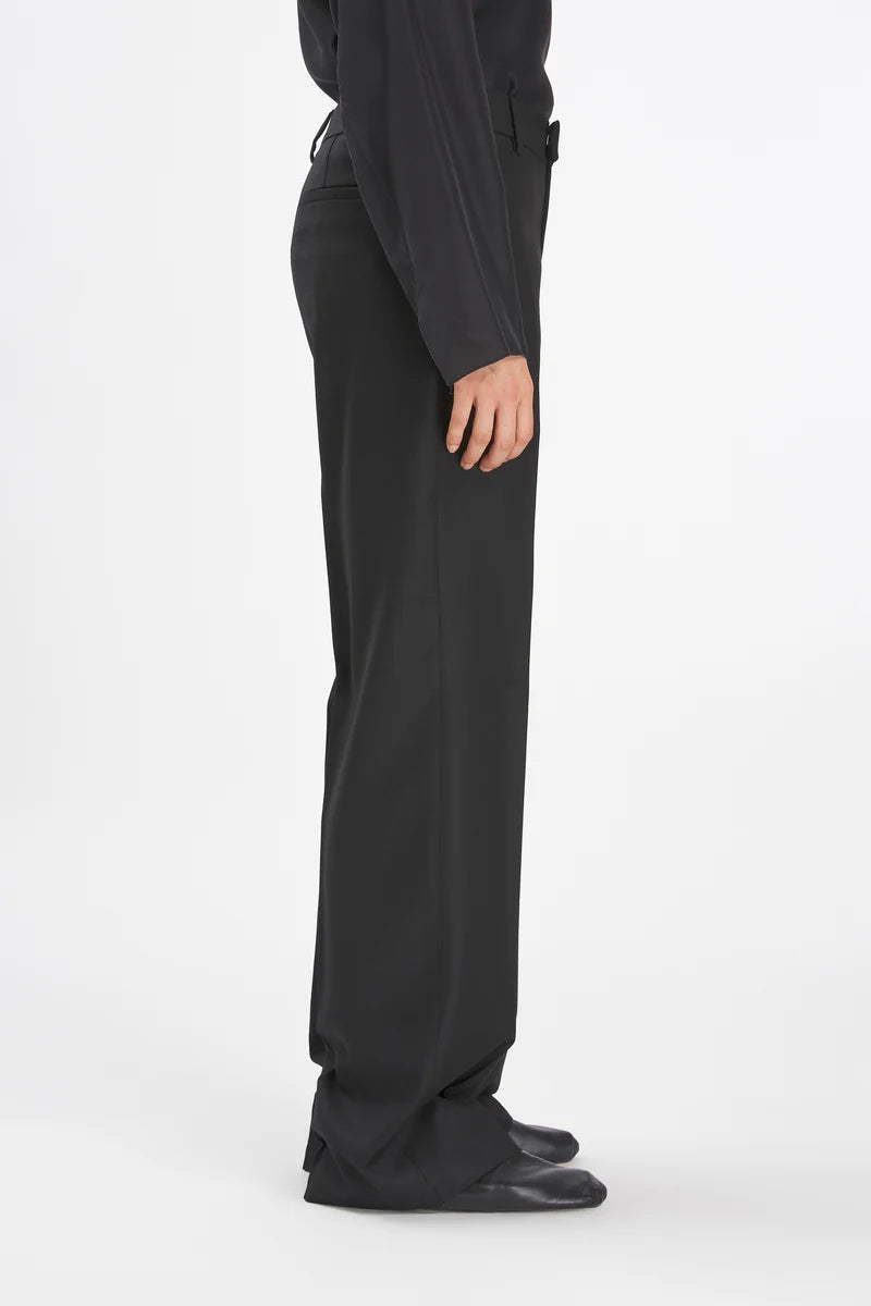 A side profile view of a person wearing black wide-leg trousers, a black long-sleeve top, and black shoes against a plain white studio background.
