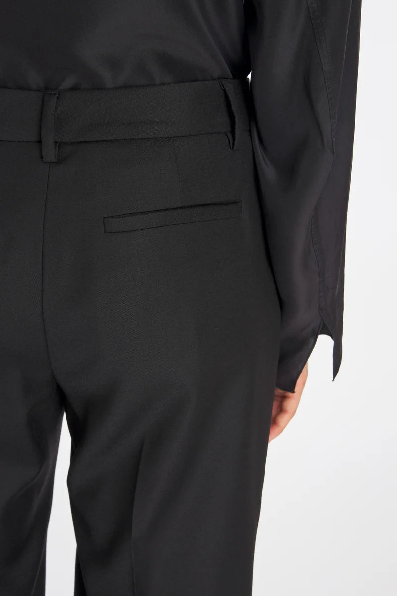 Close-up of a person wearing black tailored trousers with a belt and a side welt pocket, photographed against a plain light background.