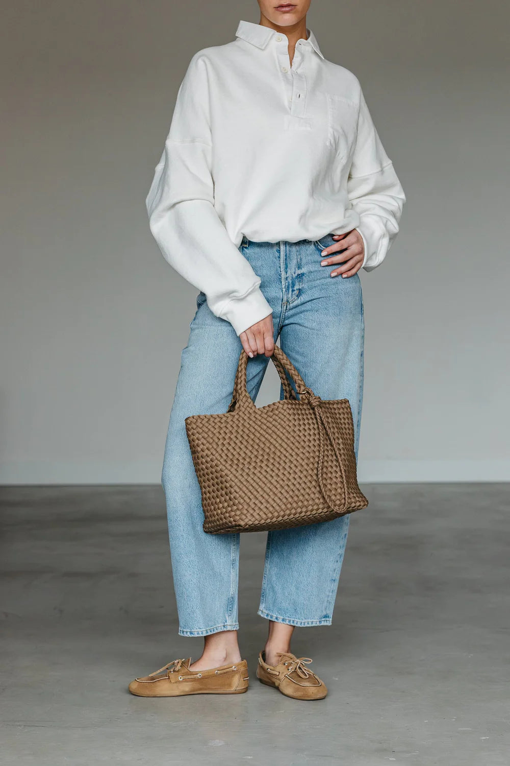 Person wearing a white shirt, blue jeans, and brown shoes holding a woven bag against a neutral background